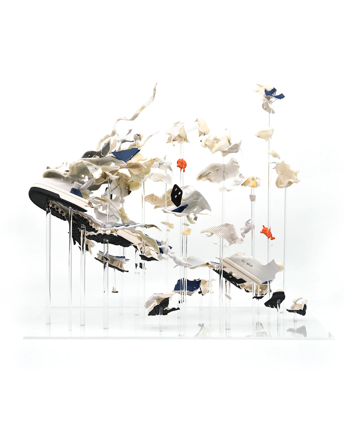 A photograph of a sculpture made from pieces of sneakers. The sculpture shows a sneaker that looks like it's exploding, with small orange figurines hanging on to various pieces.  
