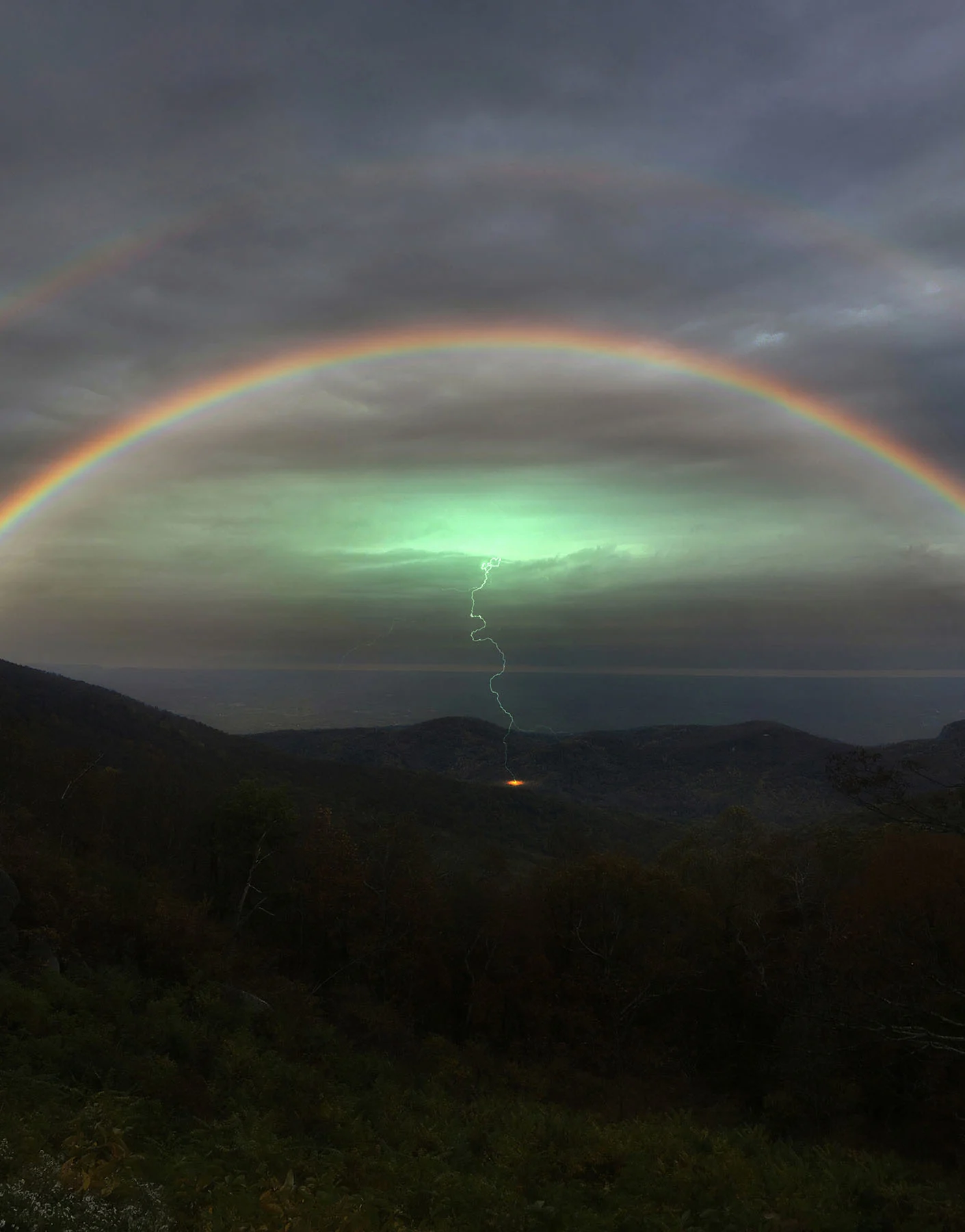 An overcast landscape photograph of a double rainbow over a lush mountainscape. At the center of the image is a single lightning bolt making contact with the earth. 