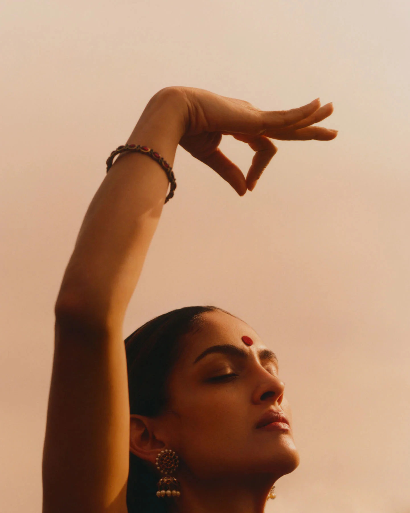 A photograph of Bharatanatyam dancer Rukmini Vijayakumar, her face softly lit by the setting sun, her right hand forming a mudra above her head, and her eyes closed in deep absorption.