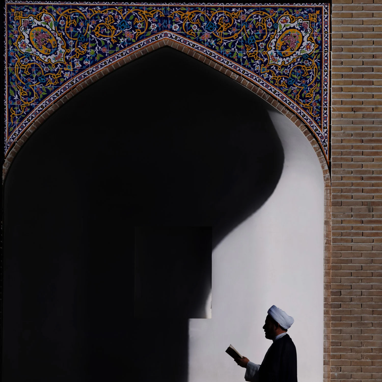 A picture of one of the clerics, featuring a man in a white turban holding one of the holy books and praying to God. Behind him sits one of the corridors of the mosque, decorated with pieces of beautiful artistic mosaics.