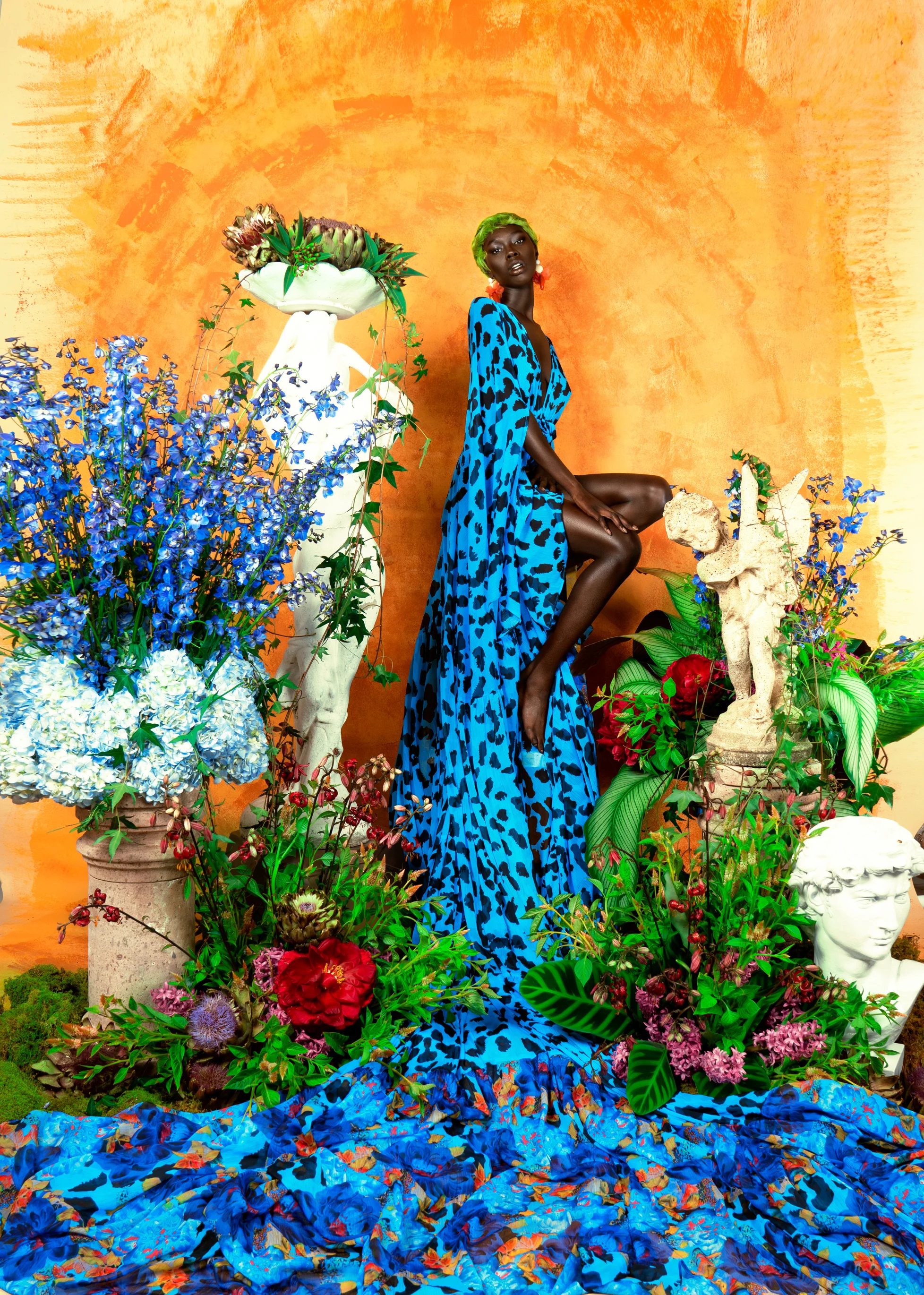 A photograph of a woman wearing a blue dress, sitting on a tall chair in front of a bright orange background and surrounded by flowers and statues. 
