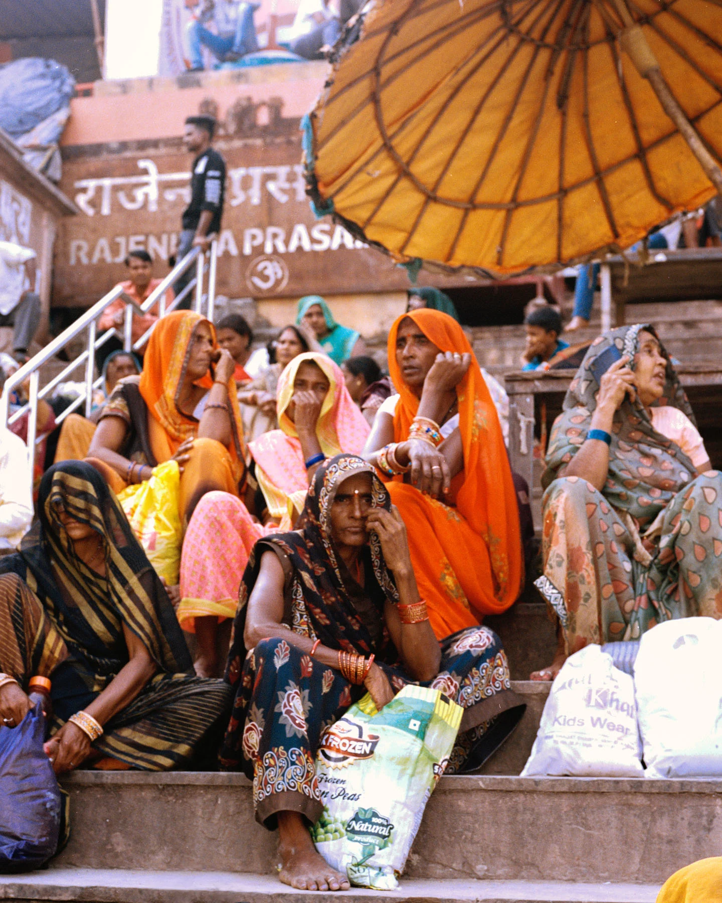 A photograph of a group of women sitting in colorful sarees on the steps of the ghat, with the loose end of the saree is pulled over their head as a facial veil, (referred as ‘ghunghat’). 