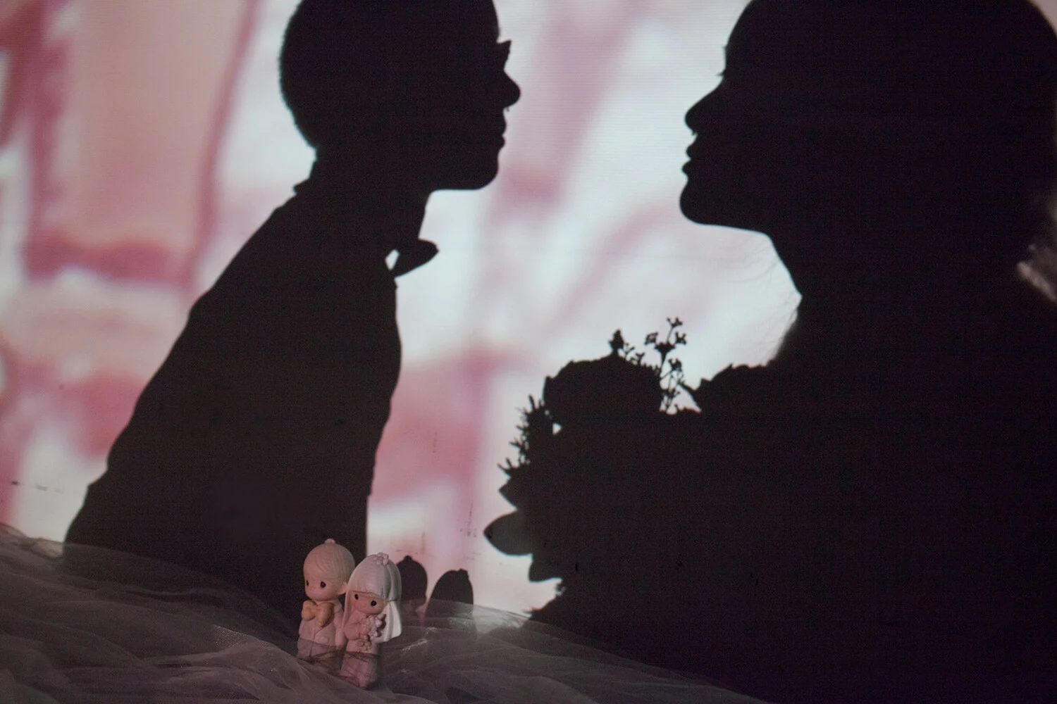 The shadows of Sy and Jonit loom over a figurine of a male-female pair in wedding attire.