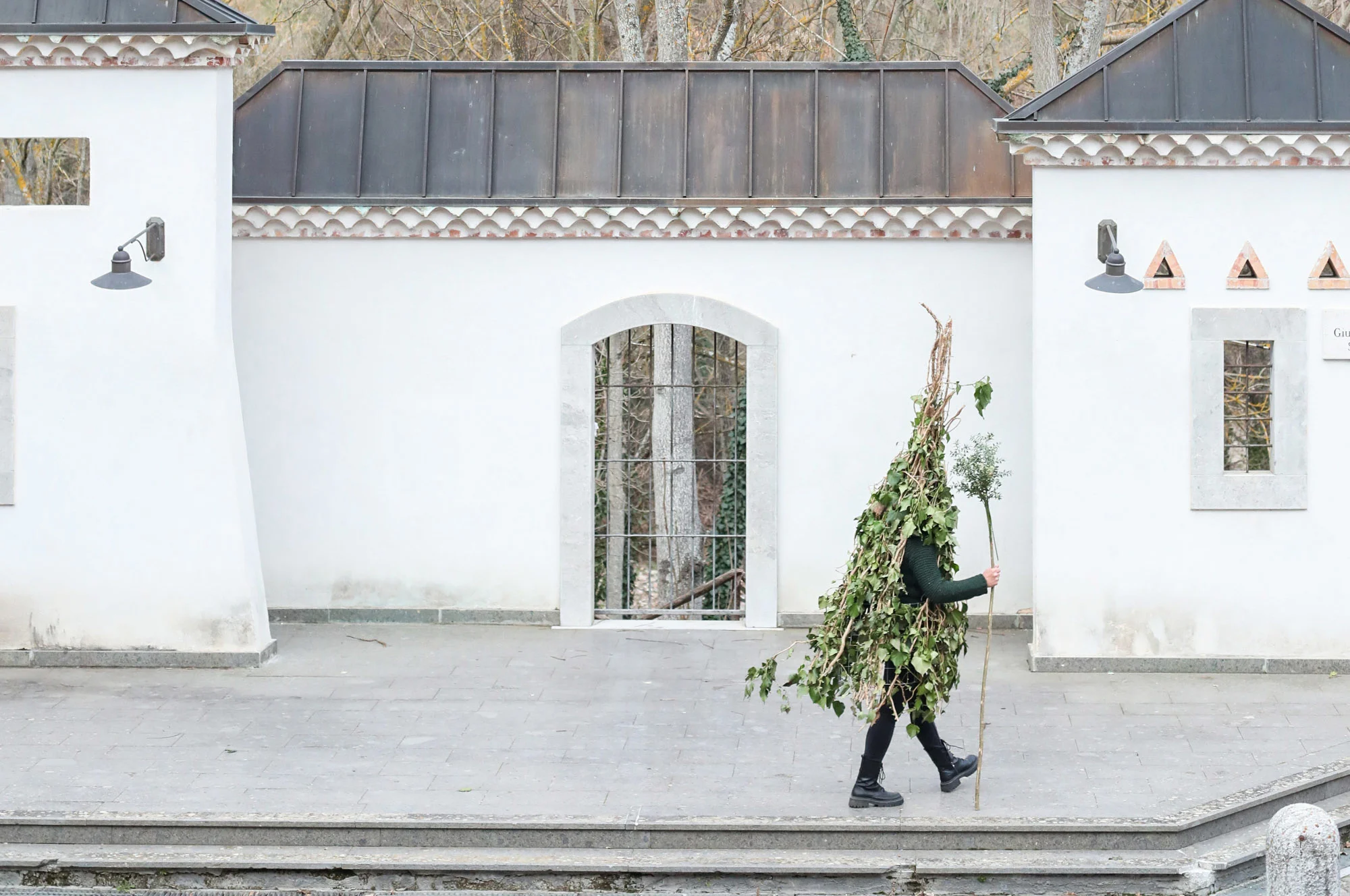 A photograph of a man walking while wearing a “Rumita” outfit (made from tree branches) through the streets of Satriano, Italy.