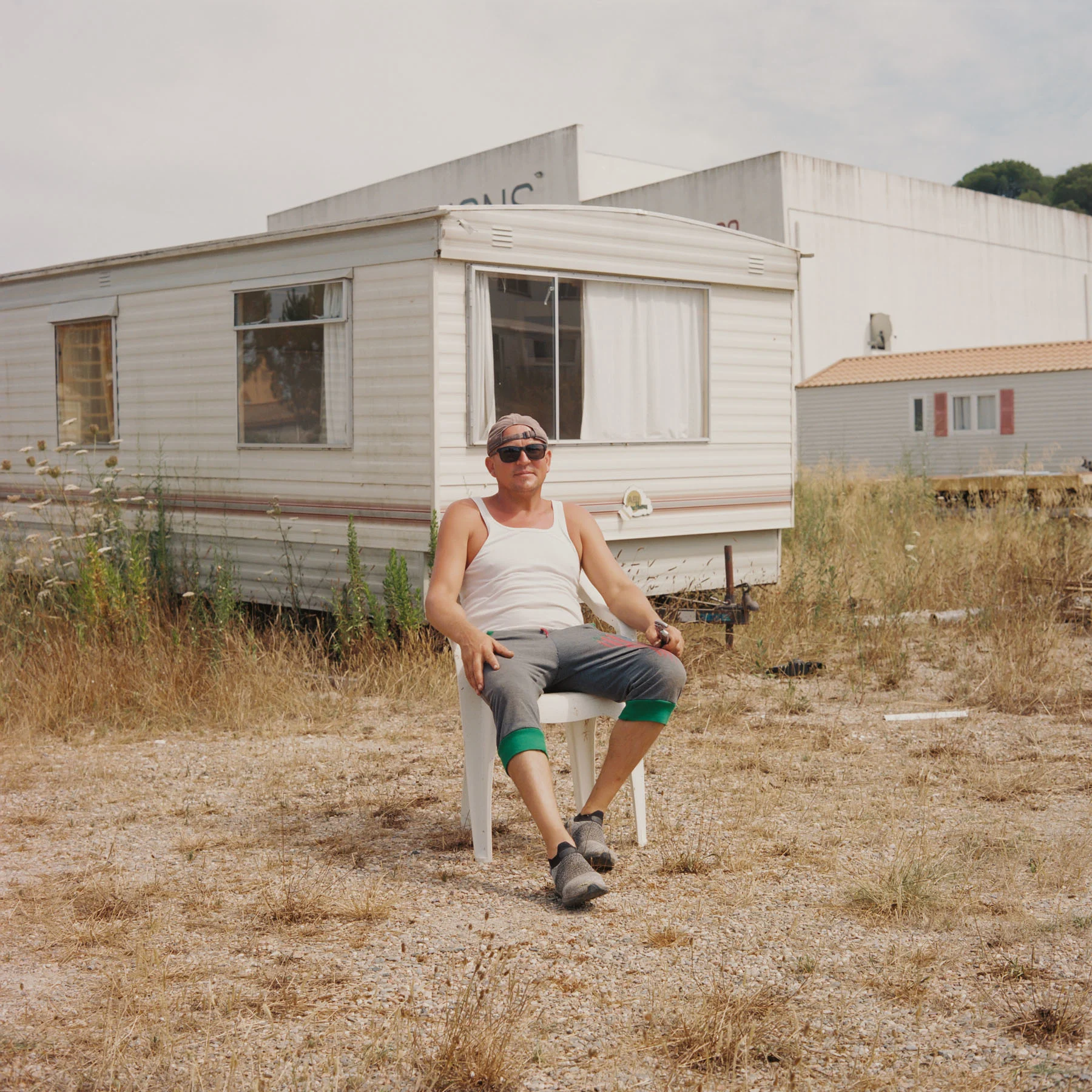 A photograph of a man sitting in a plastic white chair looking at the camera. The man is wearing pants, a white shirt and a cap. An abandoned mobile home lies in the background surrounded by white flowers and vegetation.