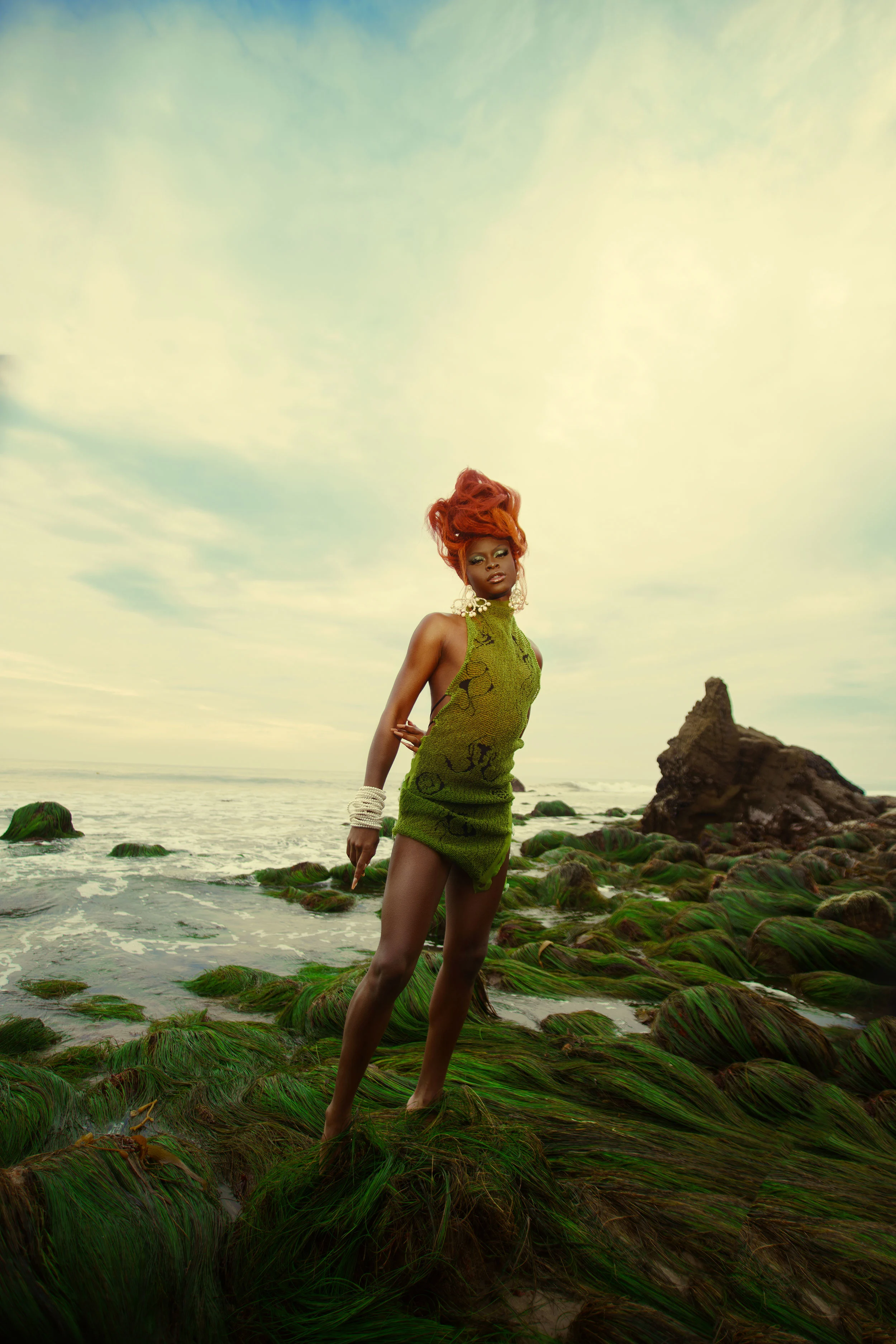 A photograph of a woman with bright orange hair and wearing a green dress, stood on some rocks next to the ocean and staring into the camera, a bright, partially cloudy blue sky above her.