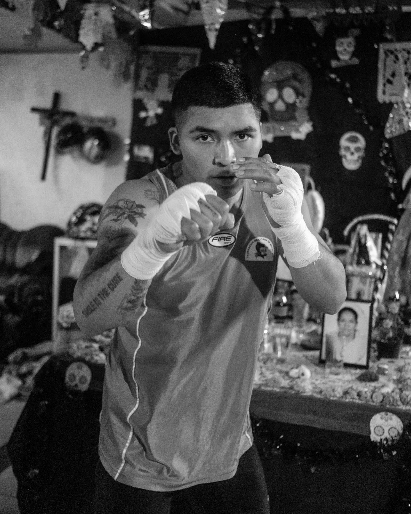 A black-and-white photograph of a boxer staring into the camera during a training session. In the background is a cross, skulls, and photos in relation to Dia de Los Muertos, a Mexican celebration honoring family and friends who have passed. 
