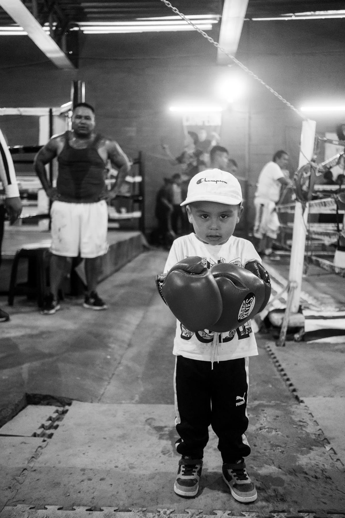 A black-and-white photograph of a small boy wearing boxing gloves in a boxing gym. His father stands behind him.