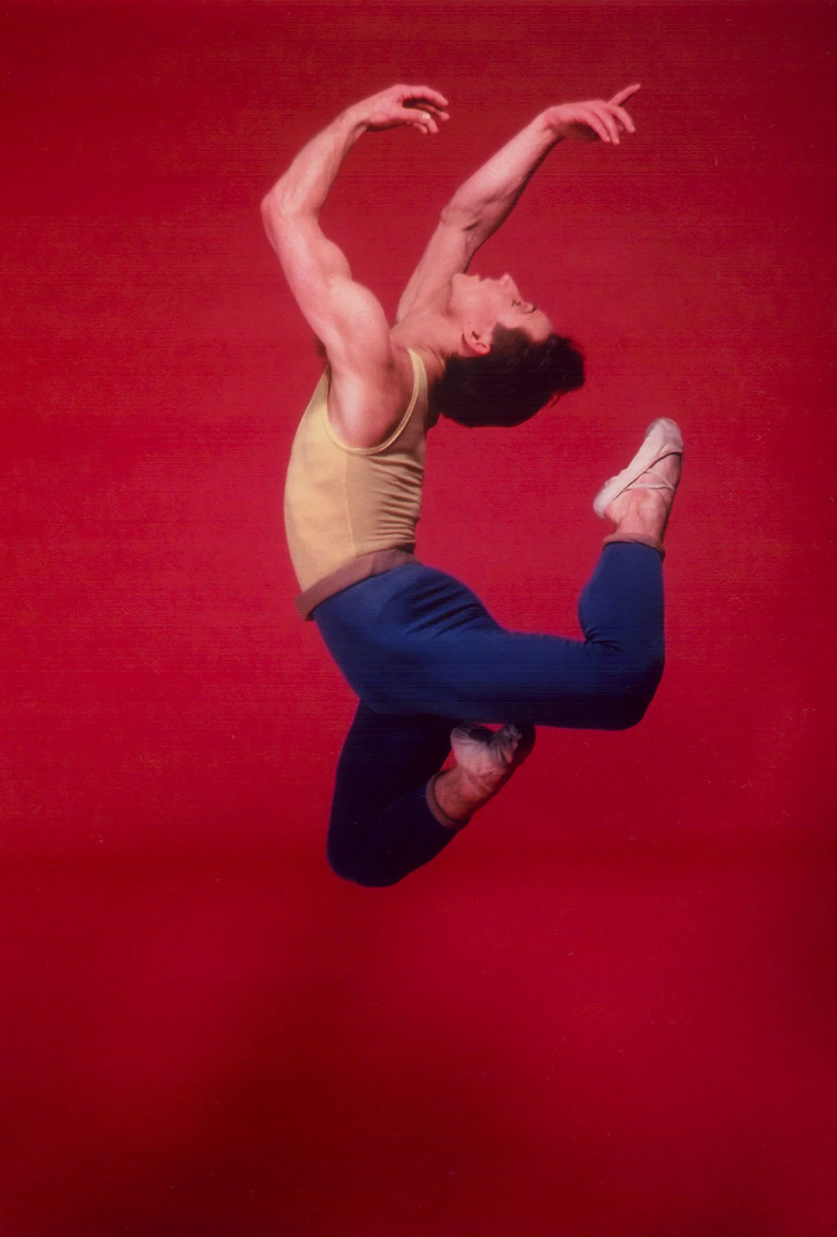 A color photograph of a male dancer caught mid-air, his arms and legs bent back. A red backdrop is visible behind him. 