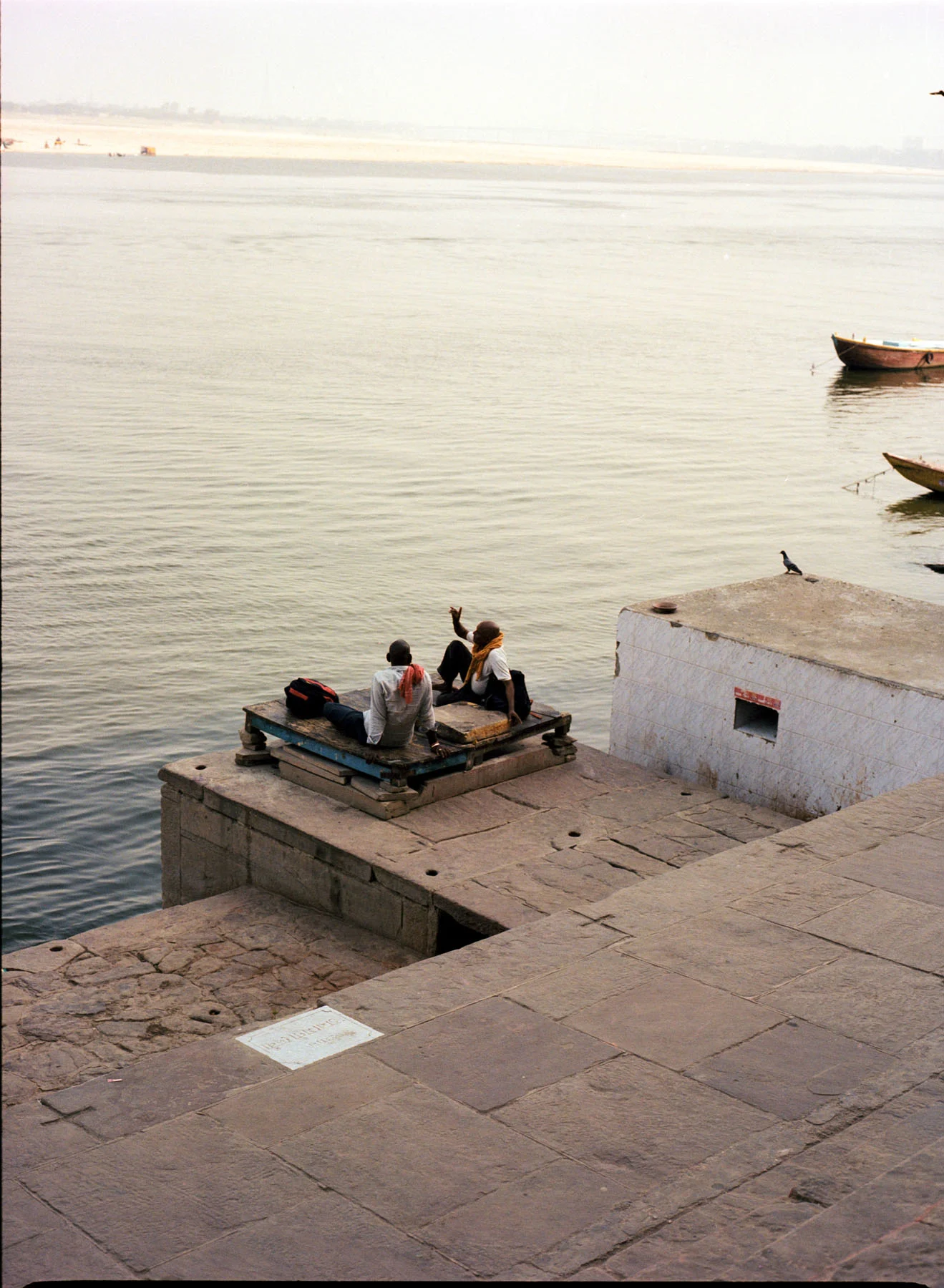 A photograph of two men sitting on the edge of the ghat (the series of steps leading down to a body of water or wharf), by the river ganges. They are chanting Sanskrit verses, with the river stretching out in the background.
