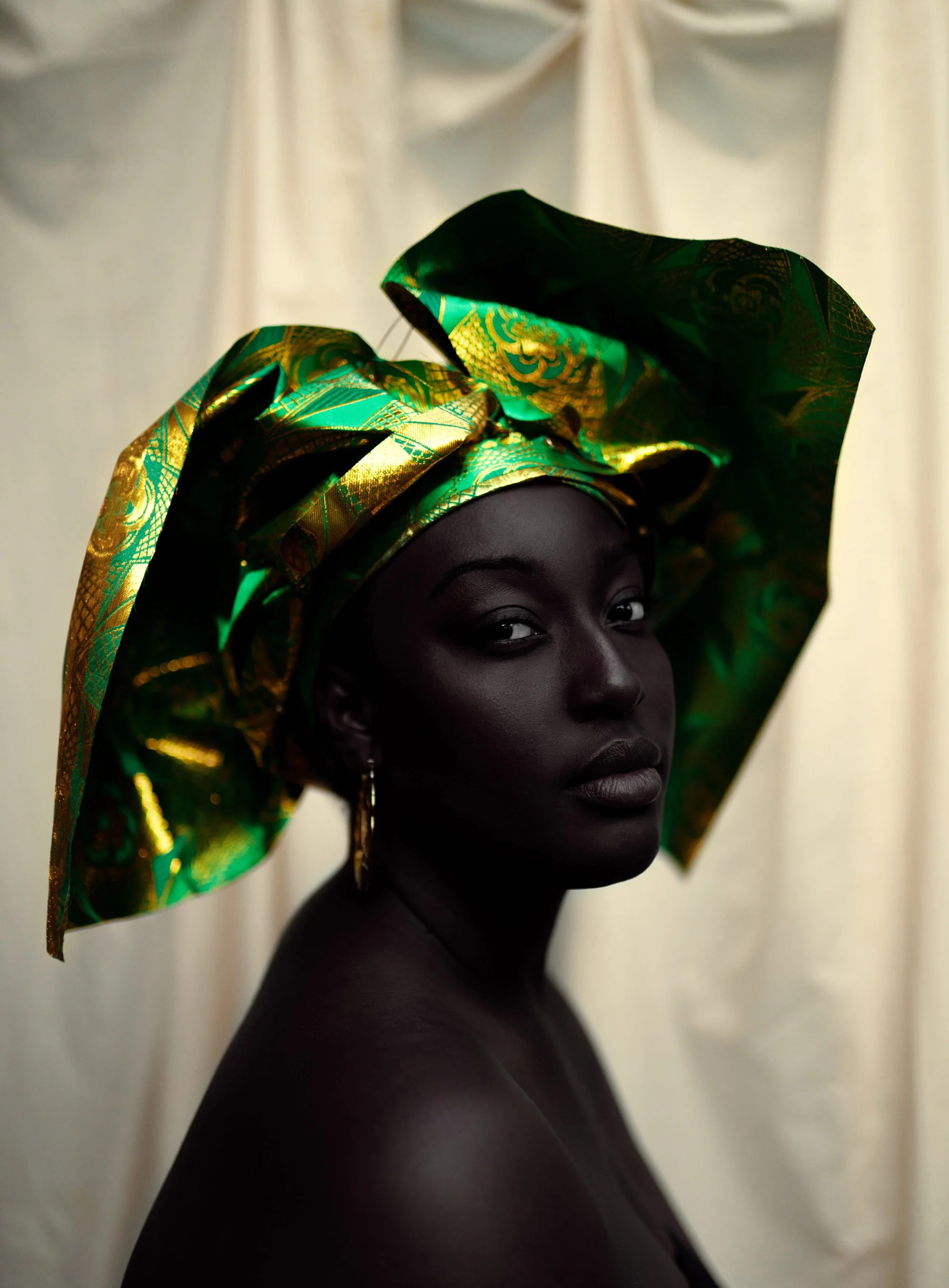 A photographic portrait of a woman wearing a green headpiece and a gold earring, sitting sideways but looking towards the camera.