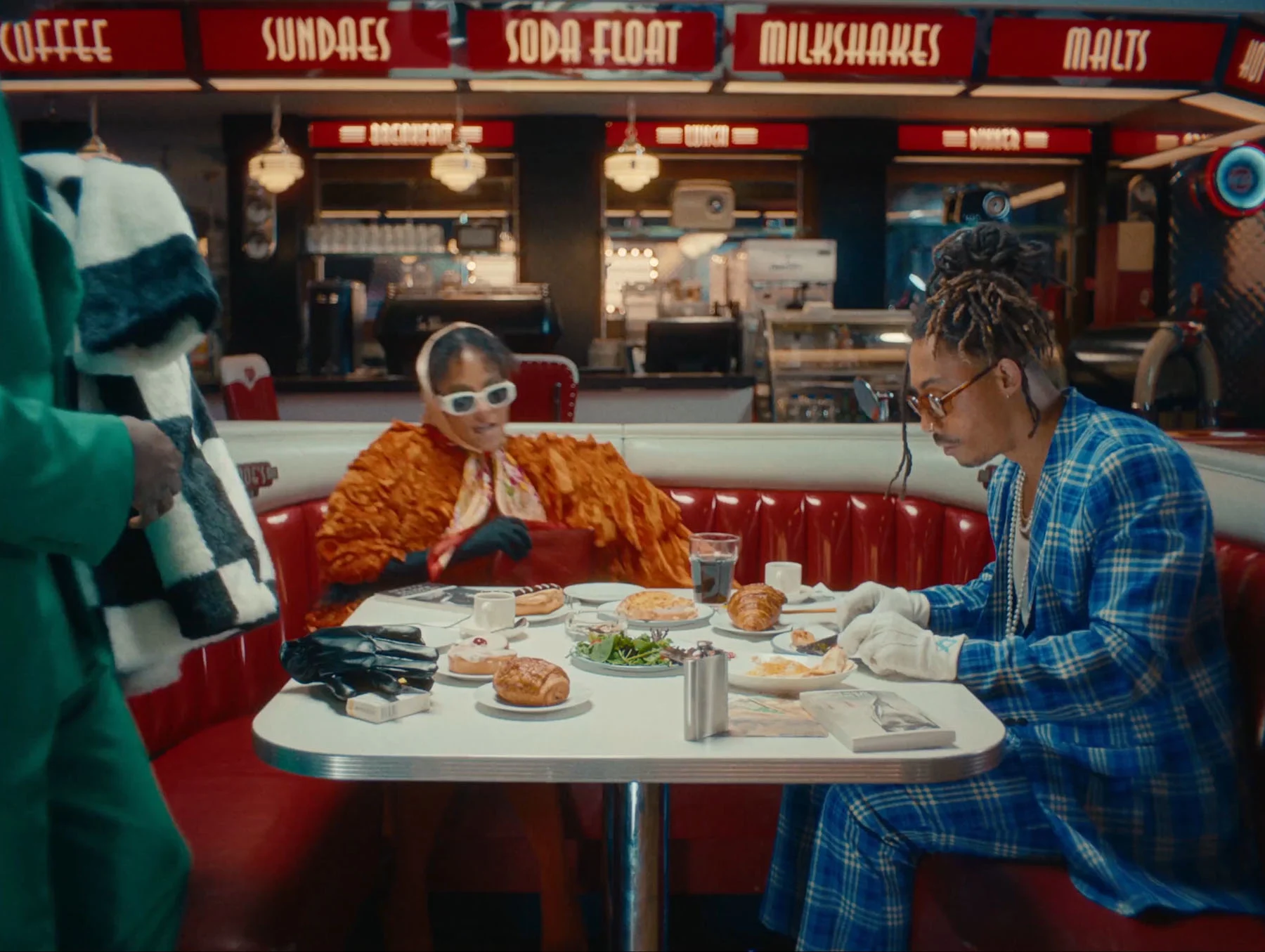 A video of three people—two men wearing suits and a woman in an orange outfit with sunglasses and a headscarf—take their seats at an American-style diner around a table filled with food and drinks.