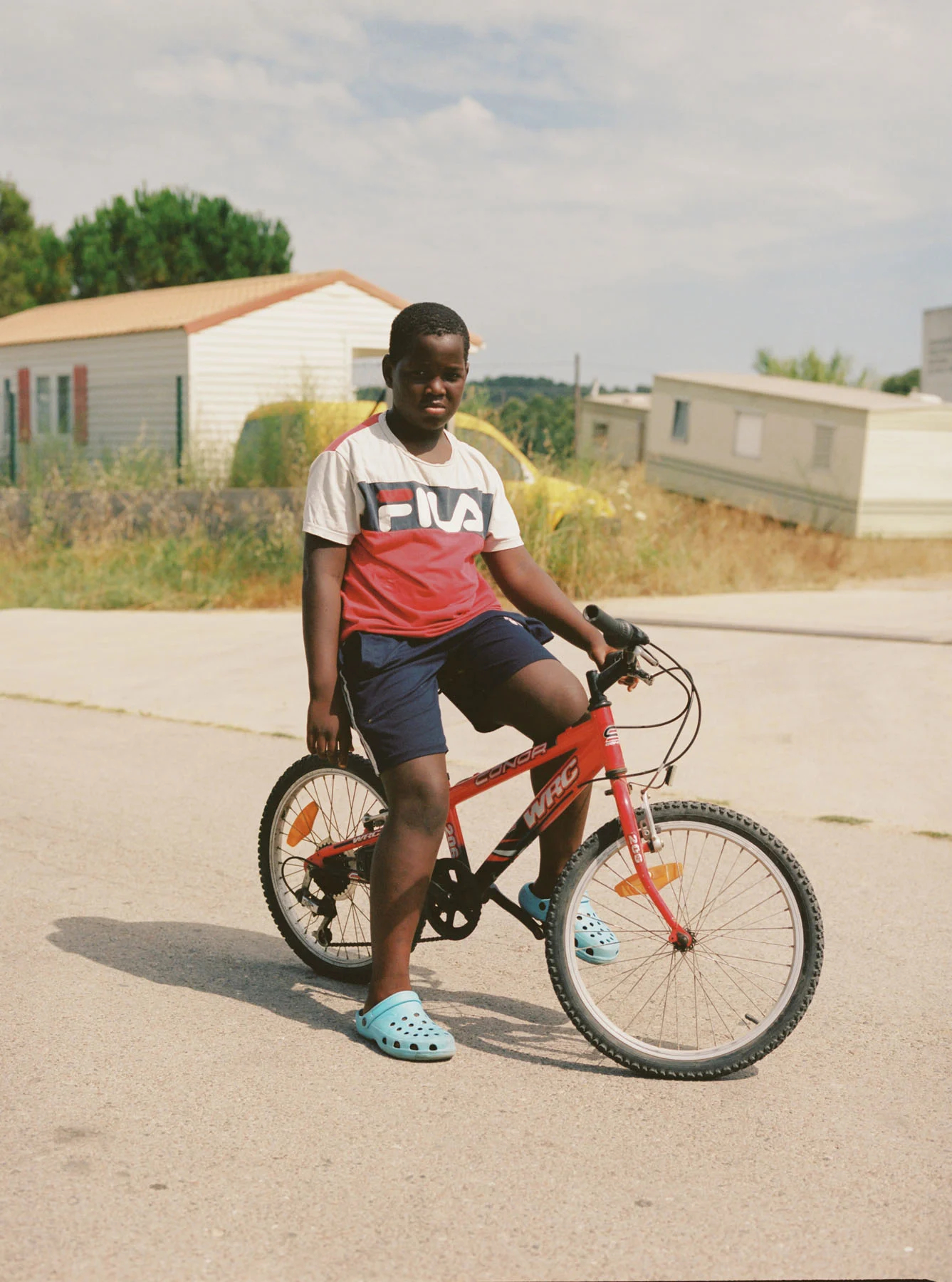 A photograph of a boy posing with his red bicycle. A few abandoned mobile homes sit in the background. 