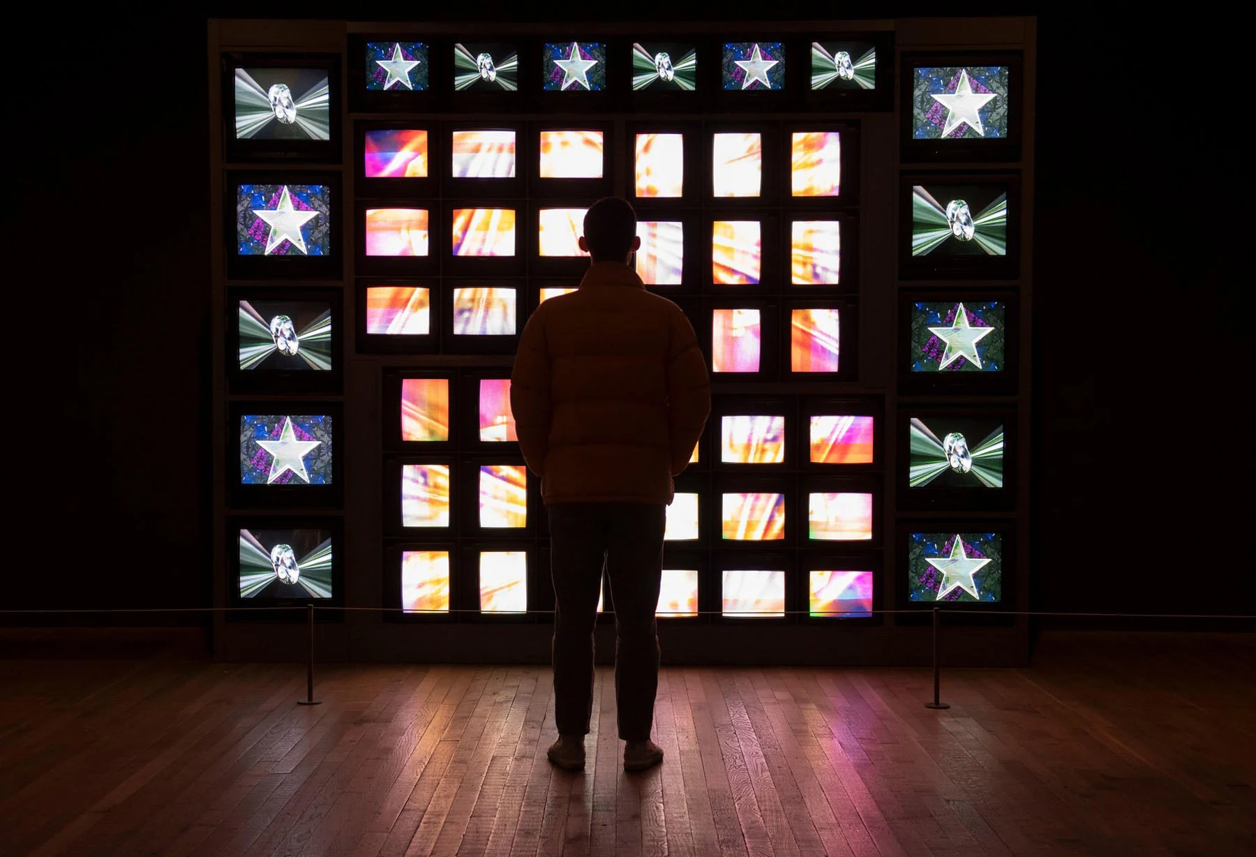 A museum visitor in a puffer jacket and trousers stands in front of a video installation. Around 50 television sets sit atop each other, playing different footage, some abstract and multicolored, others housing cinematic visuals of the cosmos.