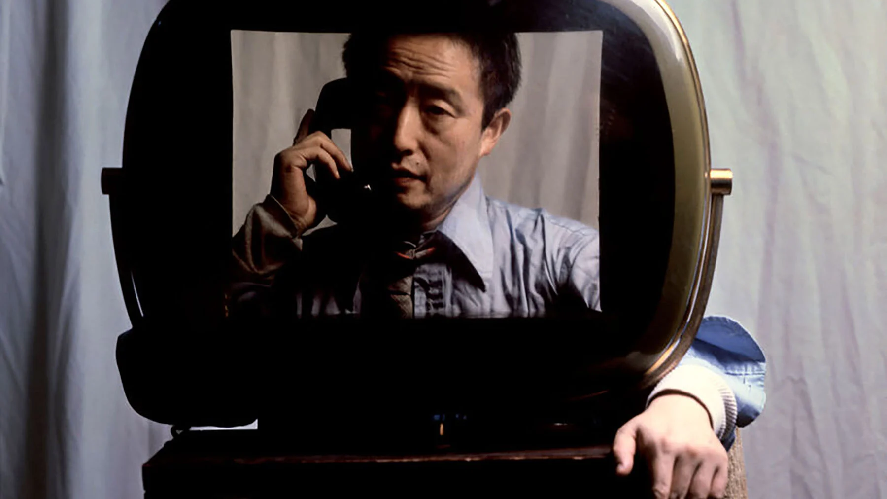 The artist Nam June Paik, an East Asian man with dark hair and wearing a blue shirt and a gray tie, is pictured on a television screen placed on a table against a blue-gray fabric backdrop. He is framed from the chest up with his face half in shadow, and he holds a telephone receiver to his right ear. The left arm of an otherwise hidden person standing behind the television screen, wearing the same blue shirt, is lined up with the on-screen image, making it appear the artist is both inside and outside of the television set.
