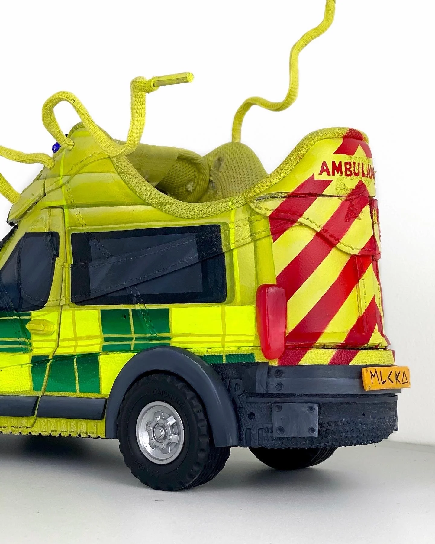A photograph of a sculpture: a sneaker that has been turned into an British ambulance, including wheels, side mirrors, number plate, lights and bumpers. 
