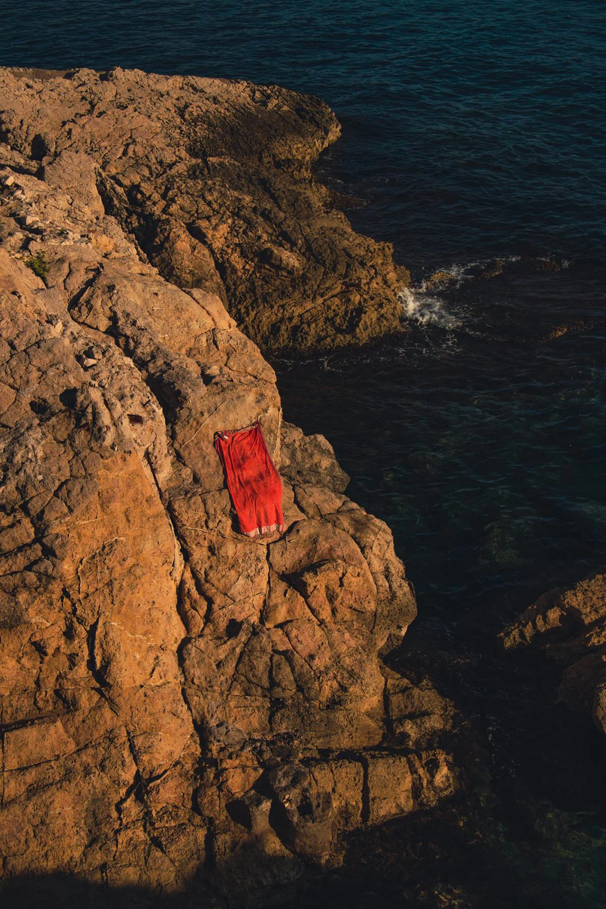 A photograph of a red beach towel lying on a rock by the ocean’s edge on a sunny summer’s day