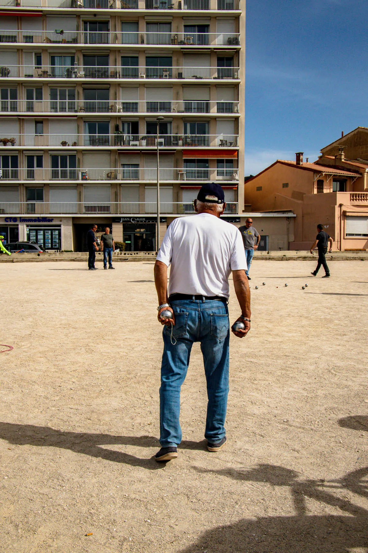 A photograph of a man wearing a white t-shirt, jeans, and a cap, playing a game of Pétanque in front of a block of apartments on a sunny day. 