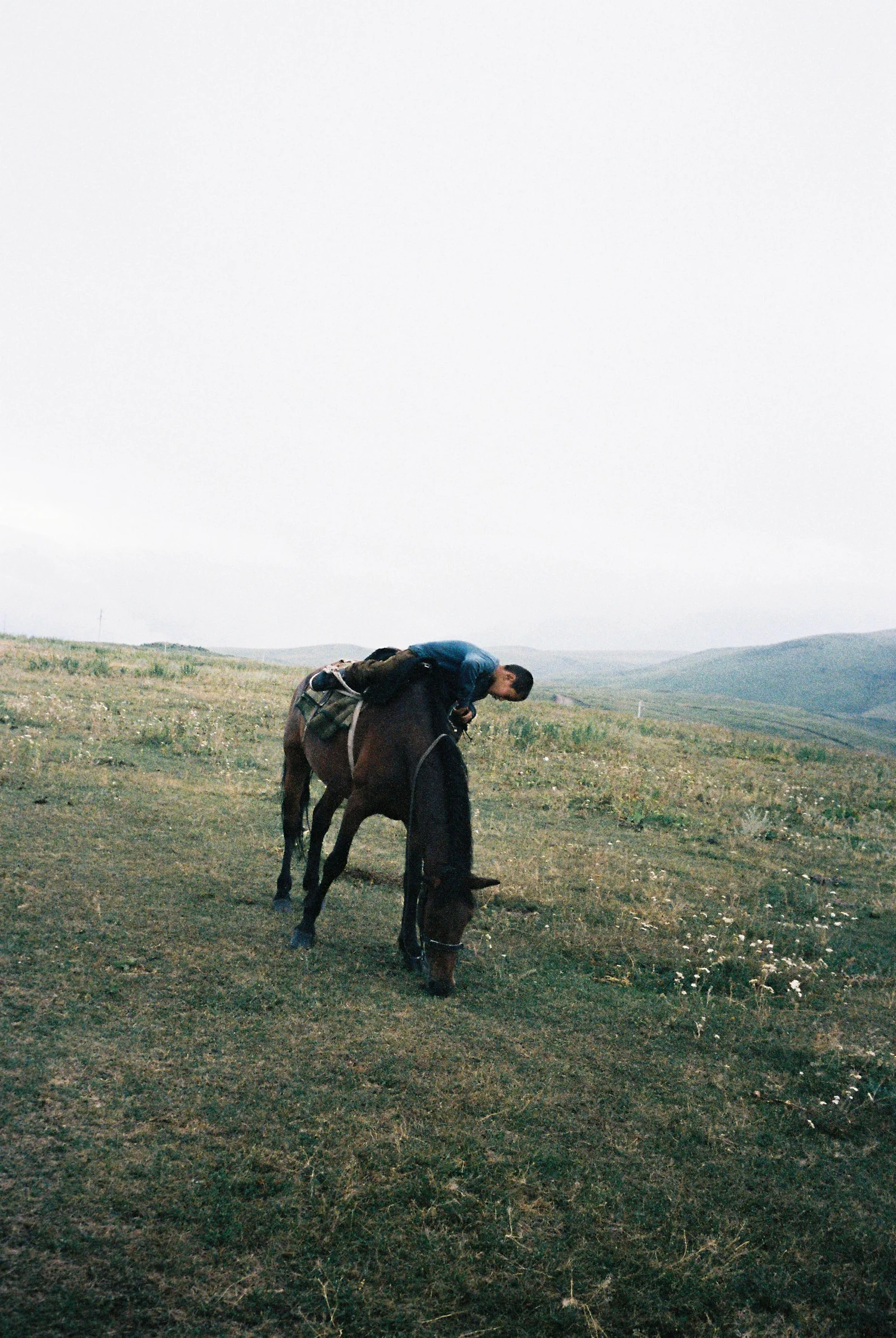 A photo of a boy on a horse in the Kazakh steppe, surrounded by mountains and fields while the horse is eating grass.