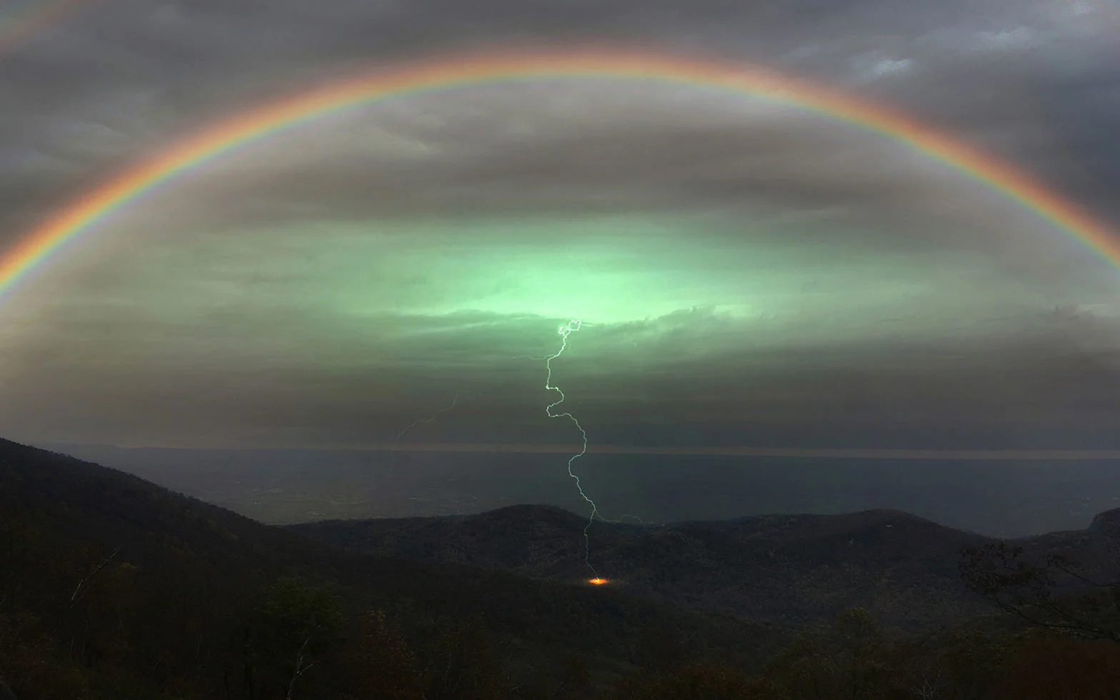 An overcast landscape photograph of a double rainbow over a lush mountainscape. At the center of the image is a single lightning bolt making contact with the earth. 