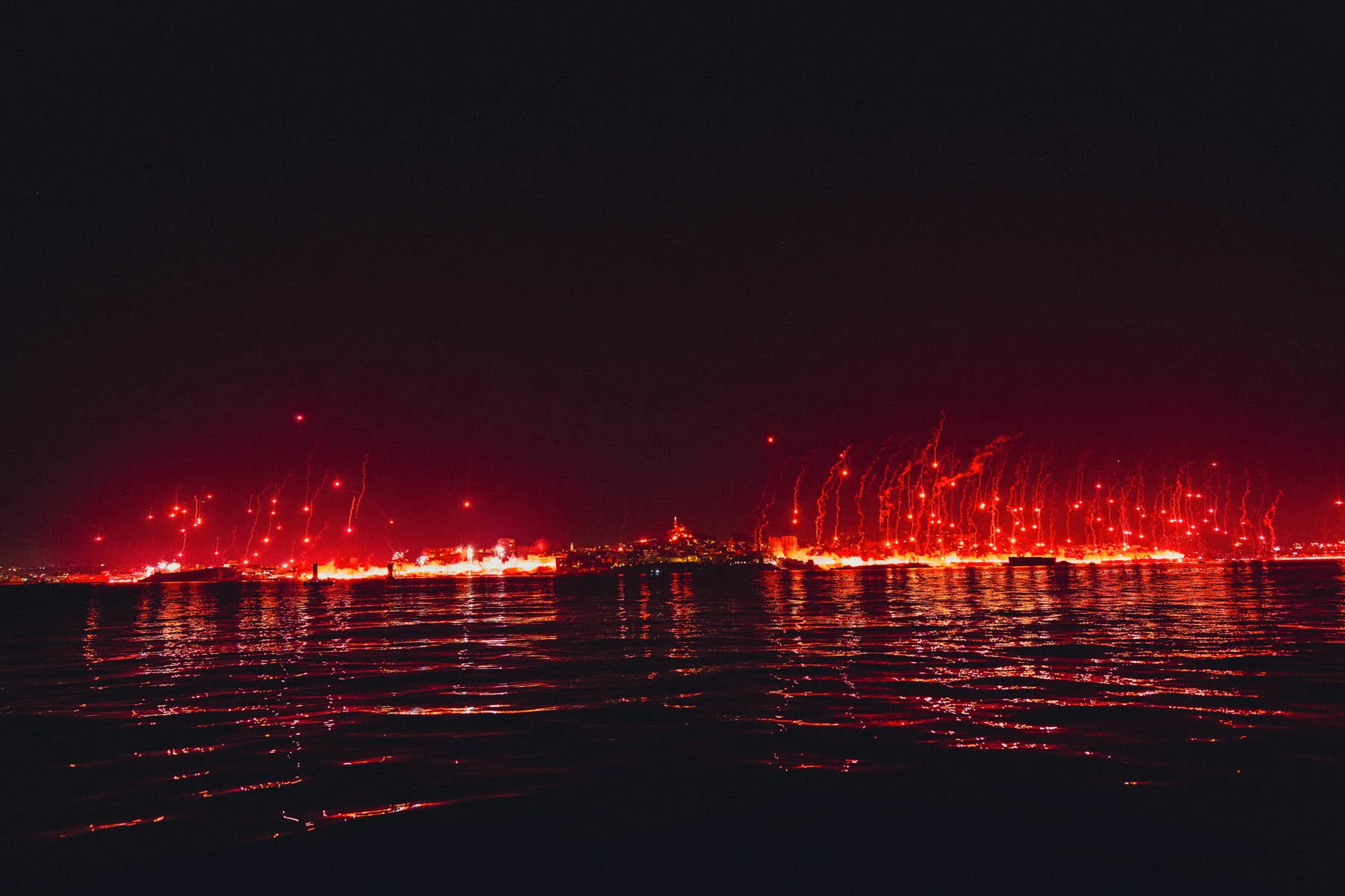 A photograph taken from a boat at night, with the waterline lit up by red fireworks and flames as part of a celebration around the anniversary of a sporting event in the field of football