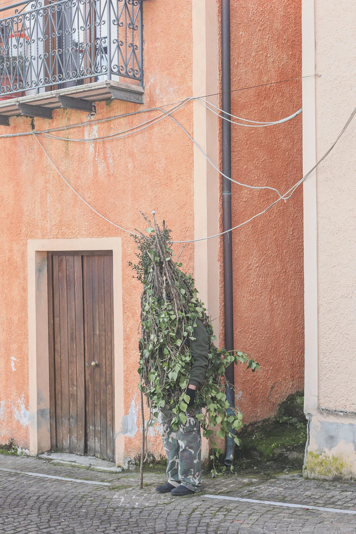 A photograph of a man dressed in a “Rumita” outfit (made from tree branches) emerging from a narrow street between two houses.