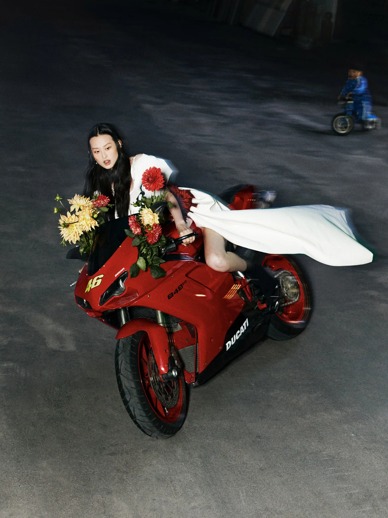 A photograph of model GuyuTong dressed in a white bridal dress as she rides a red motorcycle through a quaint alleyway.