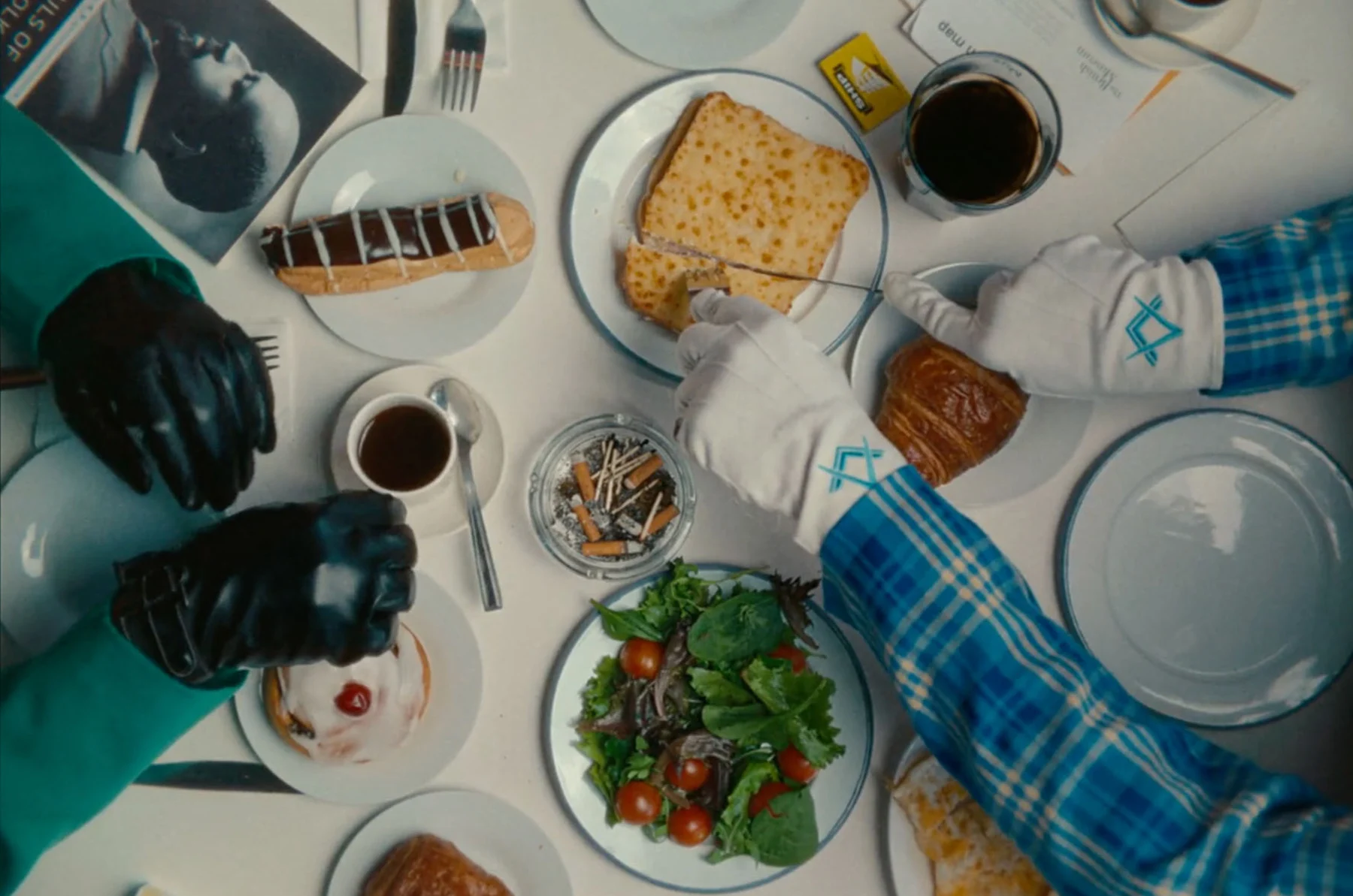 An aerial shot of a table at a restaurant. One set of hands comes in from off screen, cutting into a piece of food with a knife and fork, as another pair of hands rest on the table to the left.