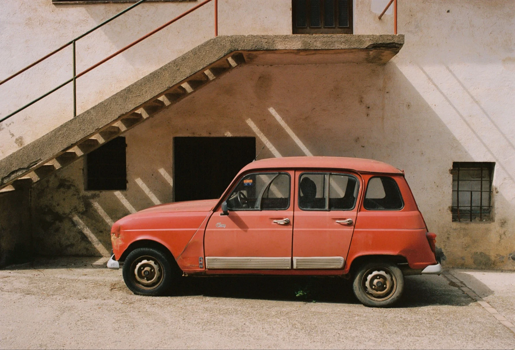 A photograph of a small and old red car parked below an old house.