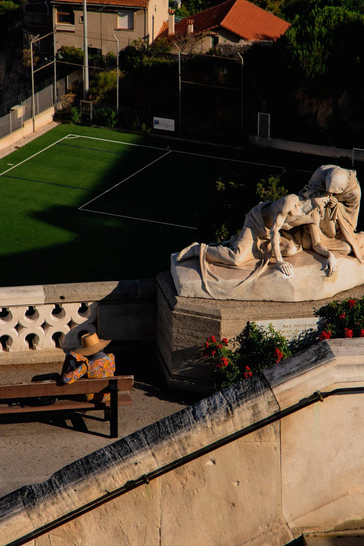 A photograph taken from a basilica with a woman wearing a straw hat and patterned long-sleeved T-shirt sitting on a bench next to a statue of Jesus and Veronica by the sculptor Auguste Carli facing a football pitch