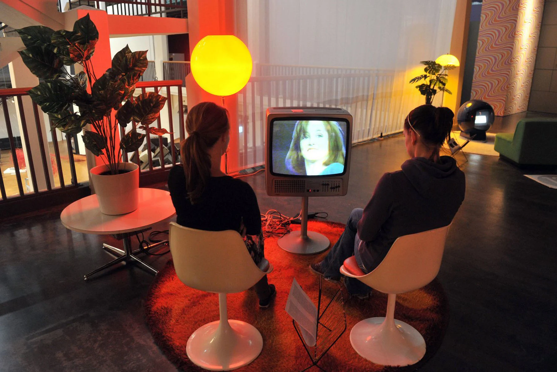 Two women sit side by side in white chairs, watching someone on the television in front of them. Around them are potted plants and furniture reminiscent of the 1960s. The room is illuminated in a red and yellow glow.