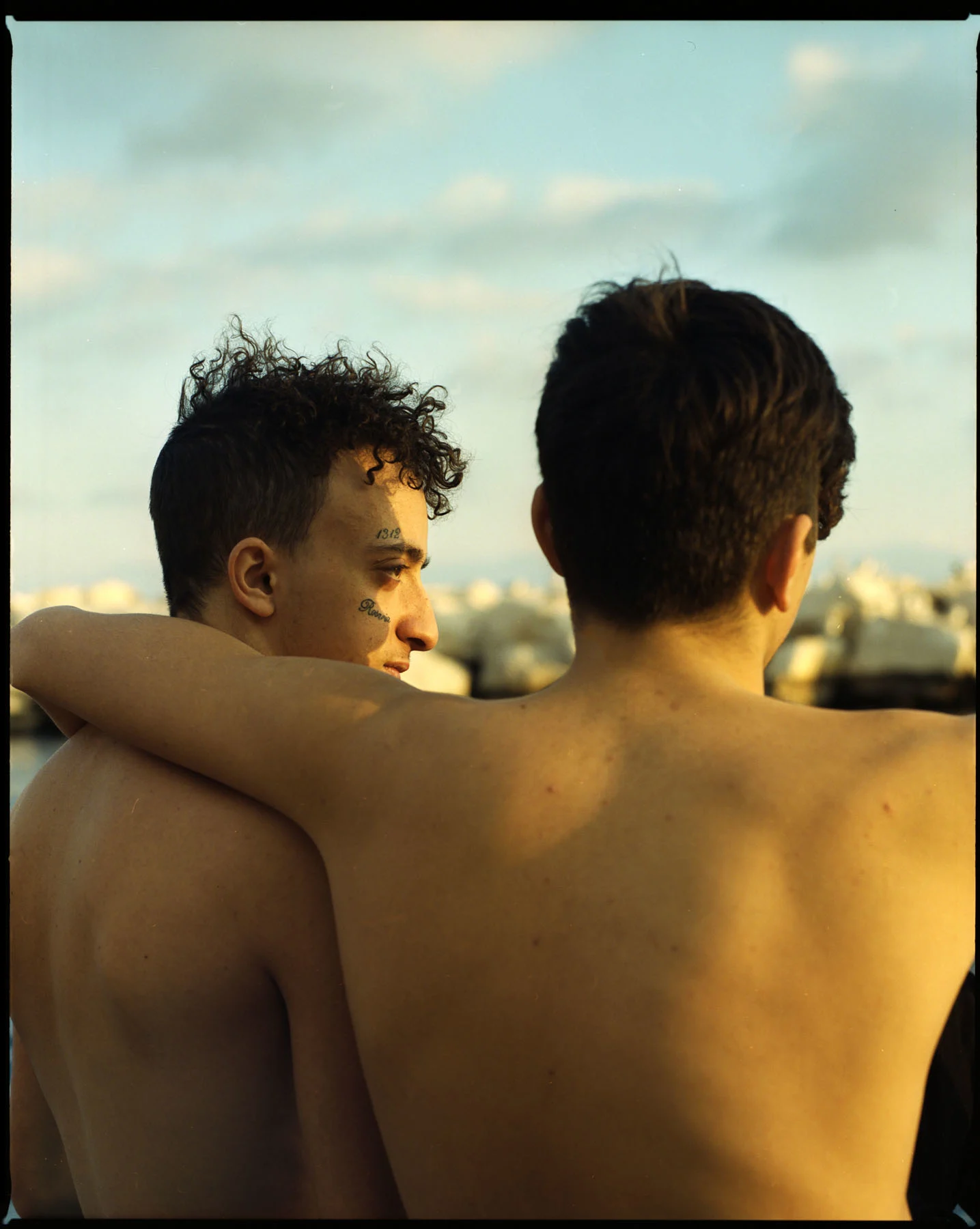 A photo of two teenage Neapolitan boys embracing. The photo is taken from behind them, the sun shining on the backs.