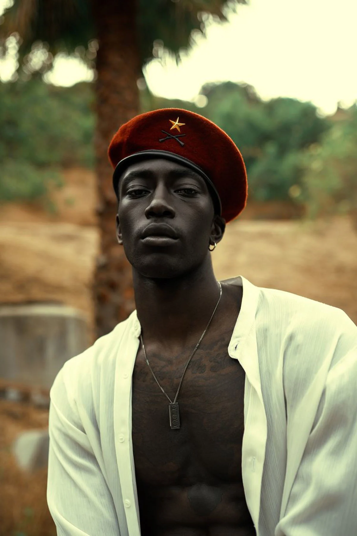A photographic portrait of a male model wearing an unbuttoned silk white shirt and a red beret with a gold star and silver chain, posing in nature, looking into the camera, his chest covered with tattoos.
