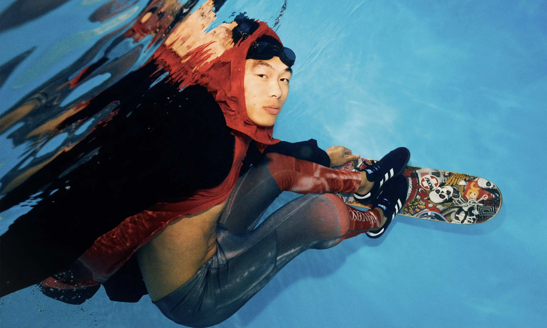 A photograph, taken below the surface of a body of water, of male model Yan performing challenging skateboard manouvers underwater.