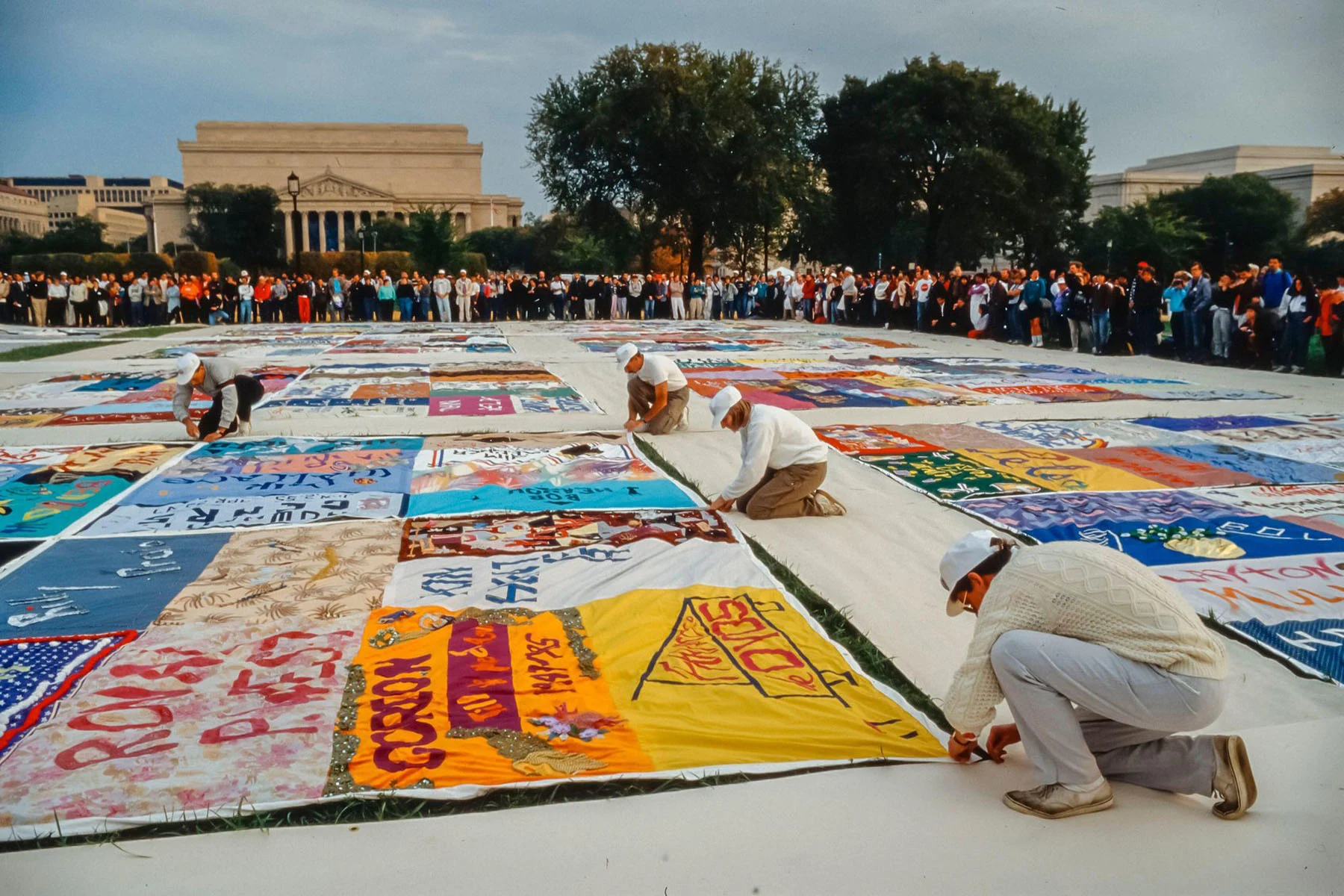 A color photograph of colorful hand-sewn quilts being laid down on the grass by four figures in the foreground. All wear white. Lines of protestors are visible behind them.