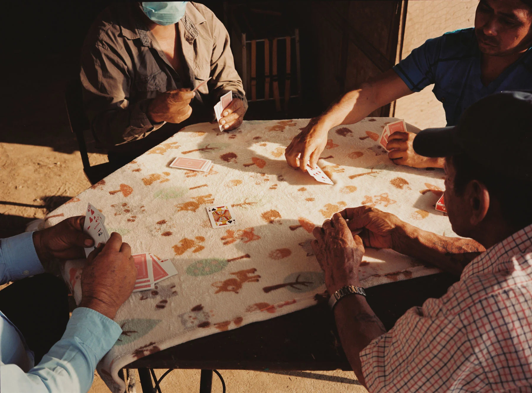 A photograph of four men playing a game of cards on a table. A warm golden light covers the scene.
