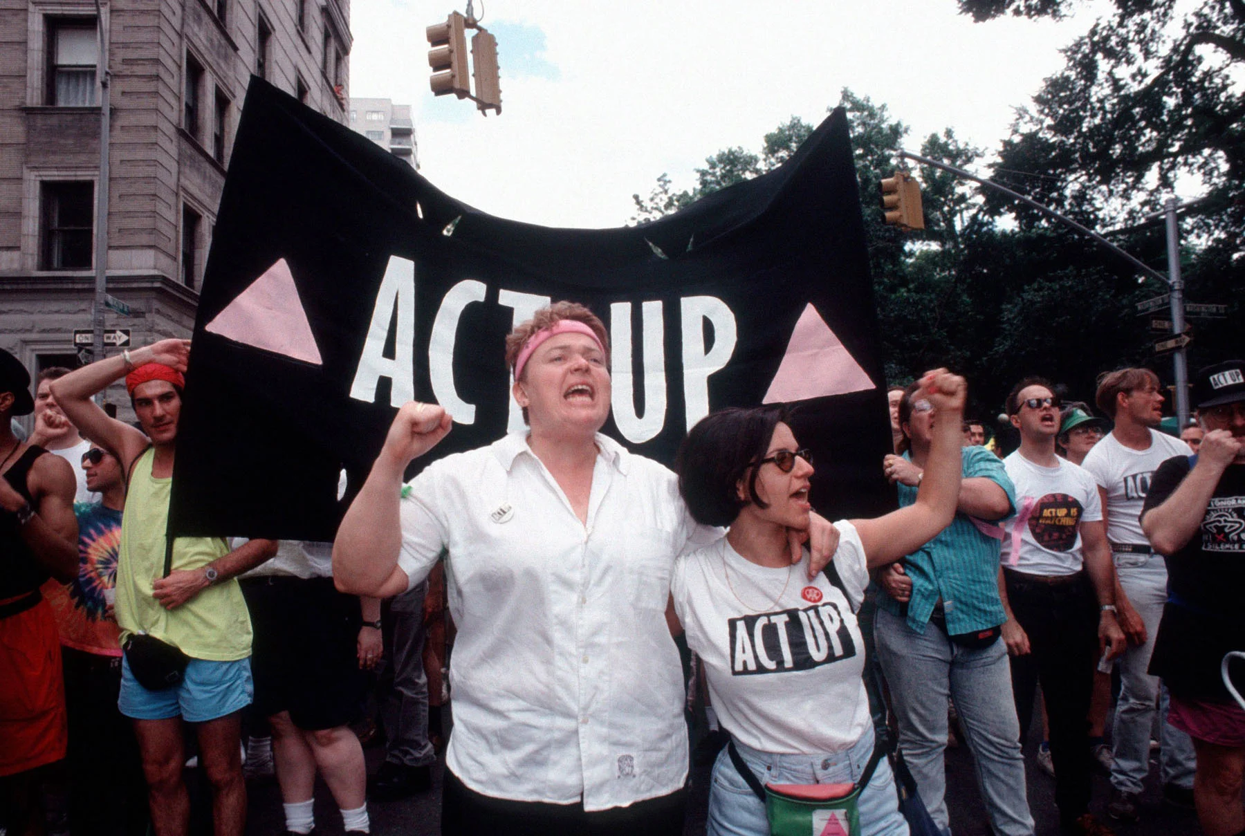 A color photograph of two people, one donning an ACT UP shirt, marching in front of a black ACT UP flag with two pink triangles on it, while a group of other protestors stands behind them. 