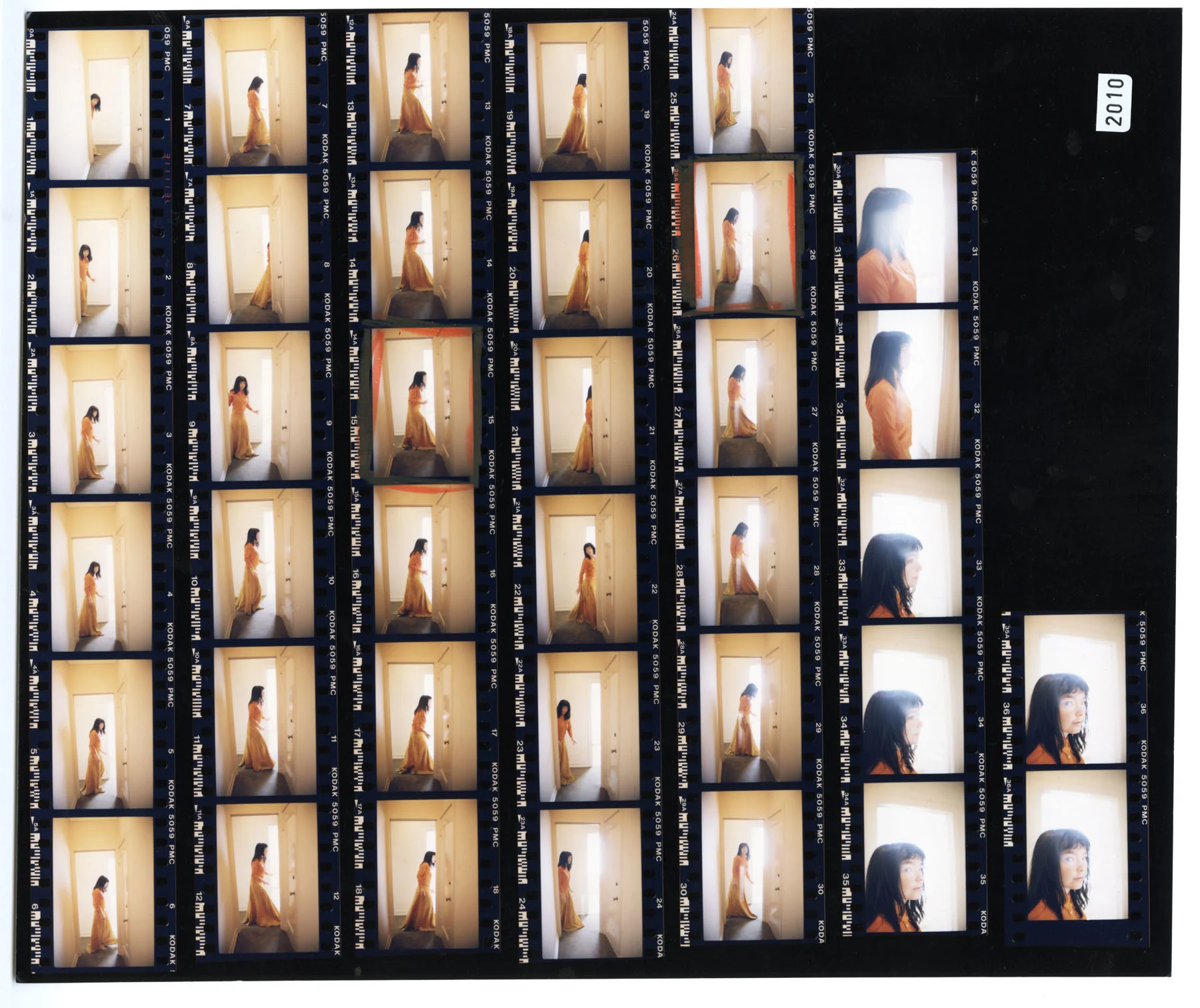 Scan of a photographic contact sheet showing multiple photographs of the musician Björk, a young, dark-haired woman dressed in an orange dress, who’s shown exploring the white hallways and rooms of a hotel.