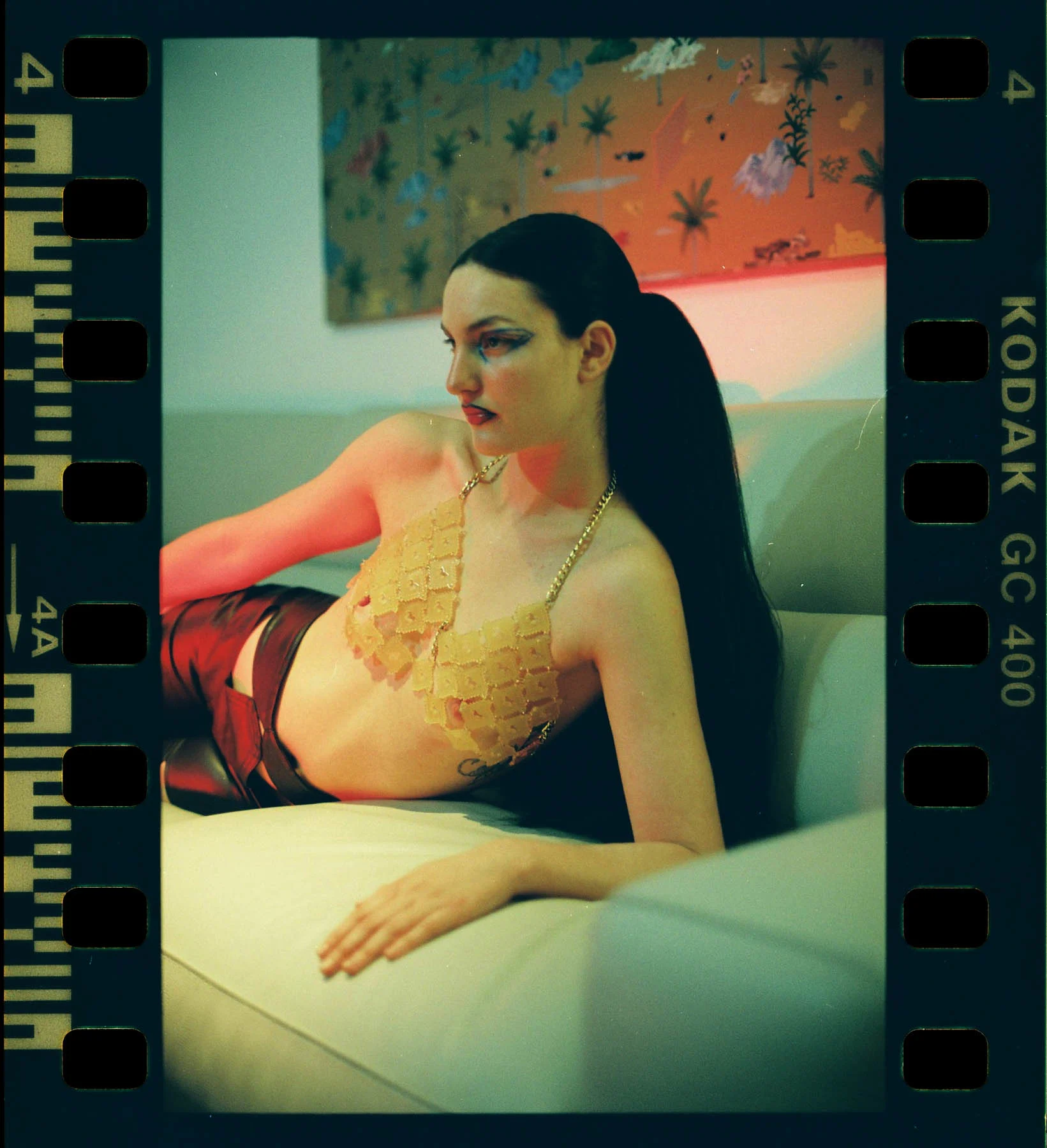 Argentinian model Carolina Vijande reclines on a couch, elegantly supporting her body with her left arm. Her long black hair is tied in a ponytail. A red light illuminates parts of her figure. The background features vibrant geometric shapes and curves.