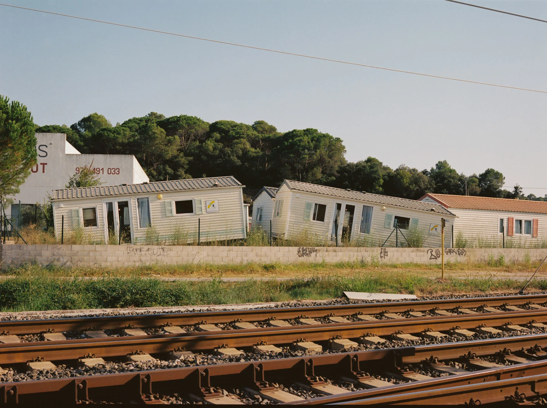 A photograph of four crooked and abandoned portable homes lying in the background between trees and vegetation. An old and rusty railroad is visible in the foreground.