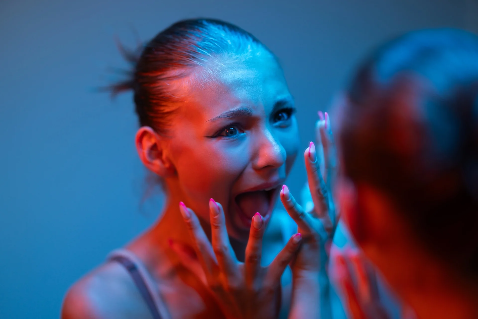 The 5 stages of psychosis concept image showing a young woman staring at her reflection, screaming and clutching her face under dramatic blue and red lighting.