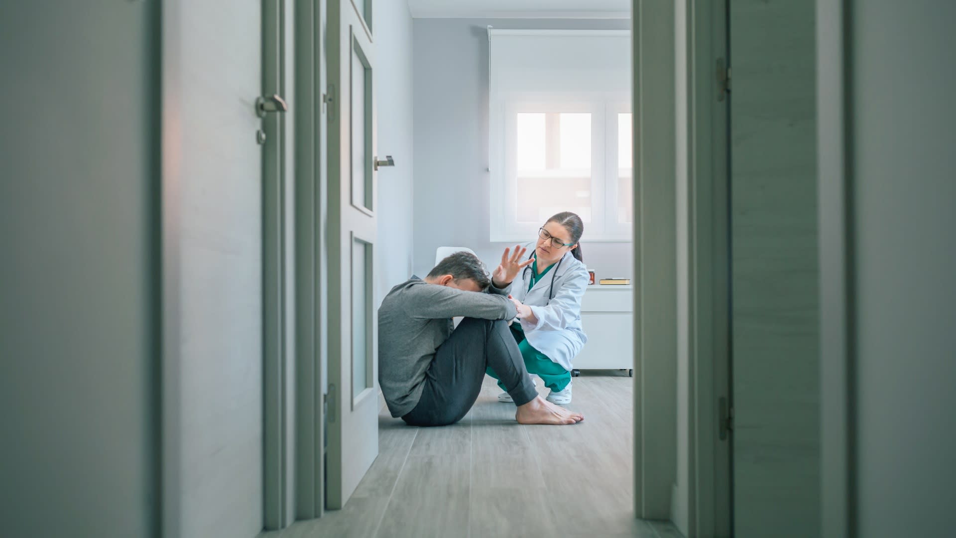 A doctor kneels beside a man sitting on the floor with his head down, offering support and reassurance during what appears to be a panic attack episode.