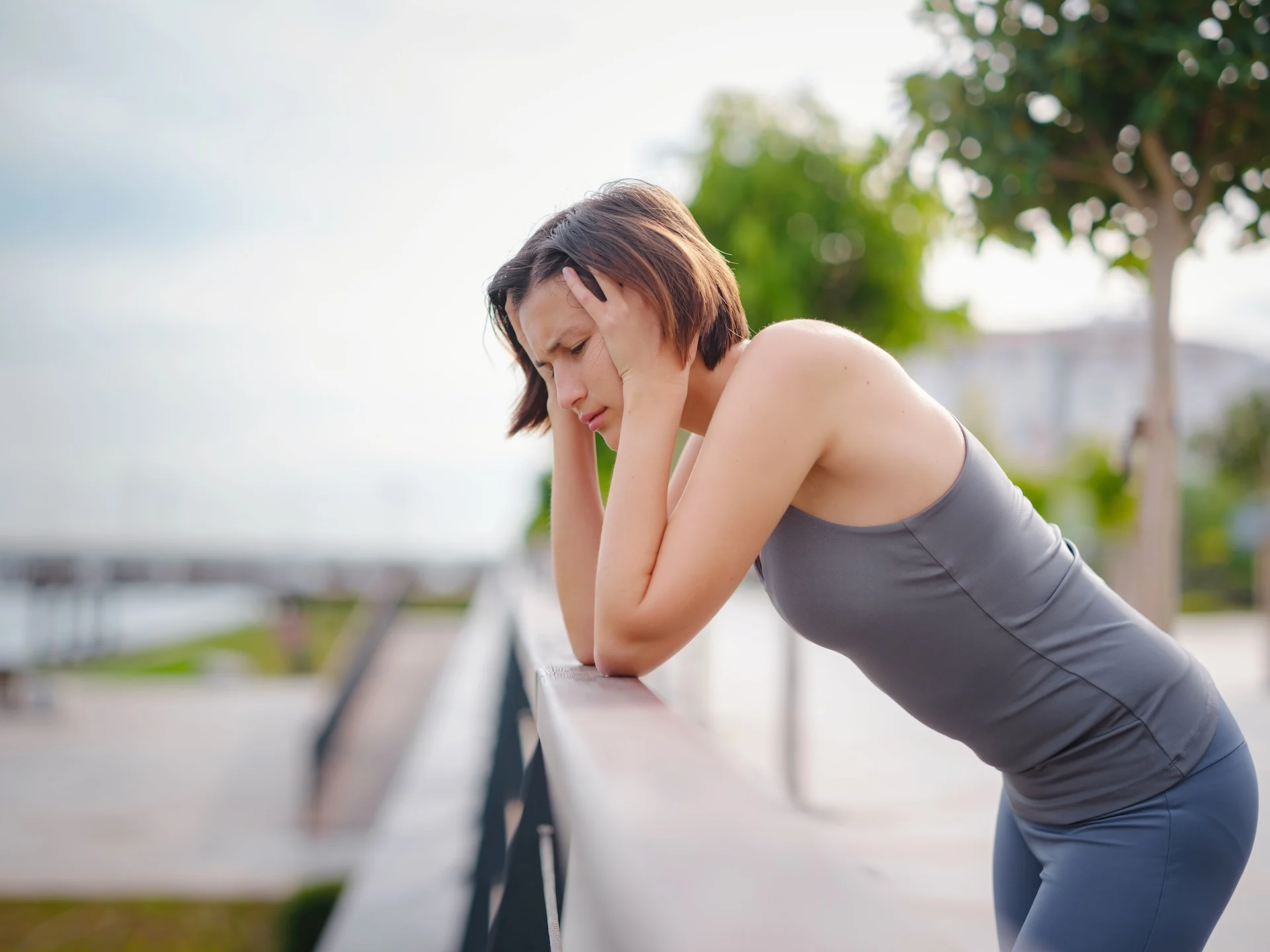What does Ativan make you feel, a young woman in athletic wear leans over an outdoor railing, holding her head in both hands and looking dizzy and distressed against a blurred park background.