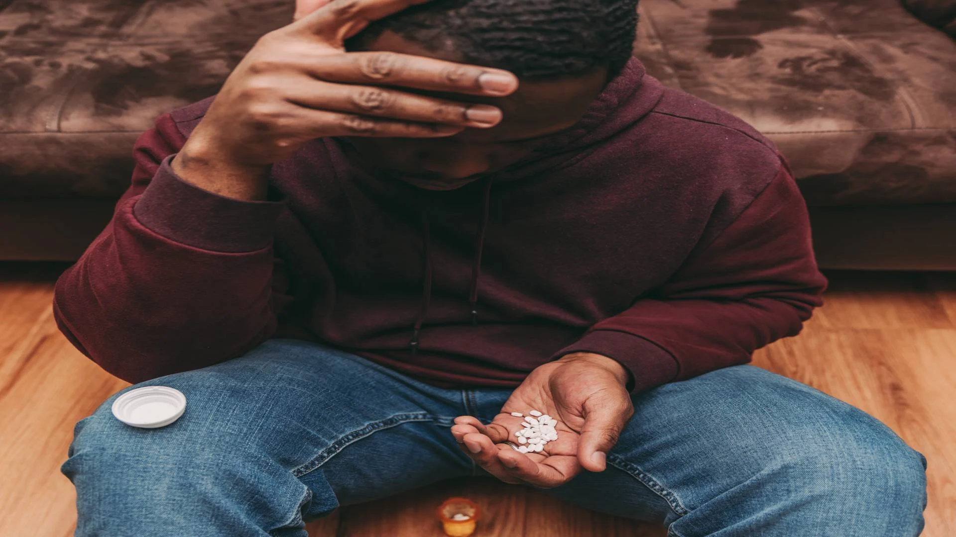 Man holding adderall pills sitting on the floor with his hand on his head in dispair