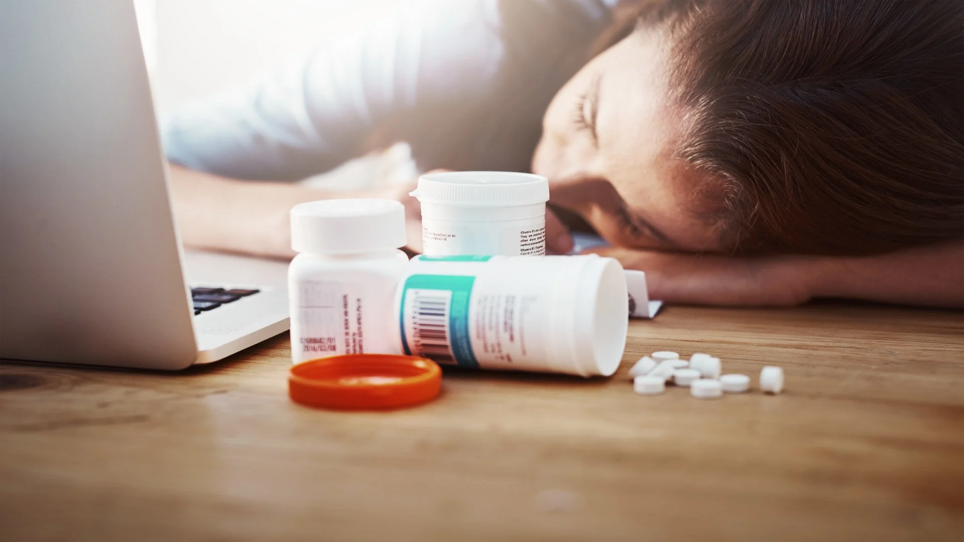 Exhausted woman collapsed on desk near medications, suggesting severe side effects or stimulant crash from Vyvanse.