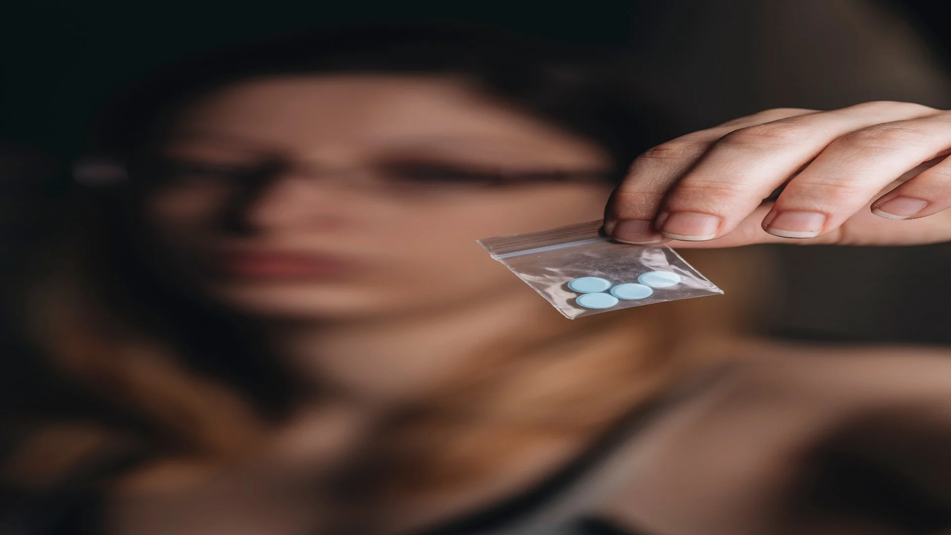 Blurry background of a woman holding a clear plastic bag containing several small blue tablets of Adderall.