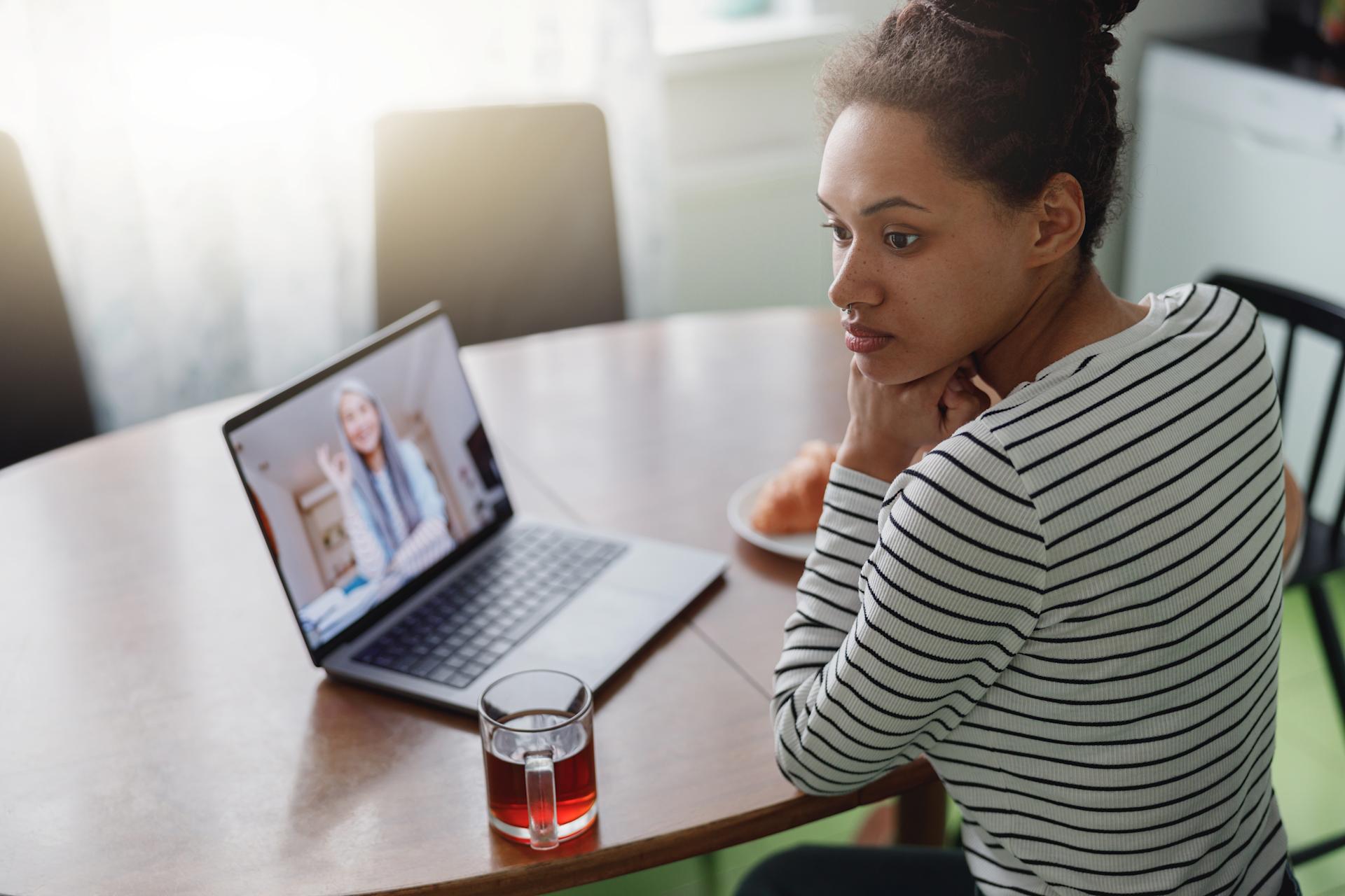 Woman participating in a telehealth addiction treatment session on her laptop, seated at a kitchen table with a glass of tea nearby.