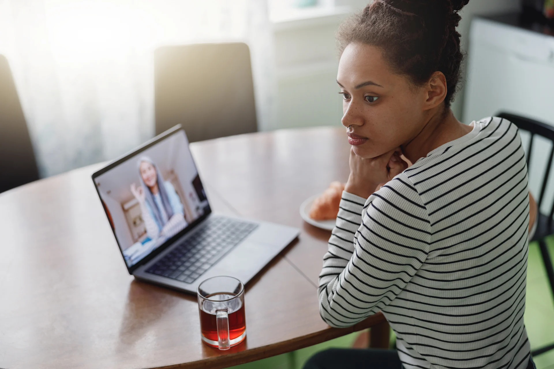 Woman participating in a telehealth addiction treatment session on her laptop, seated at a kitchen table with a glass of tea nearby.