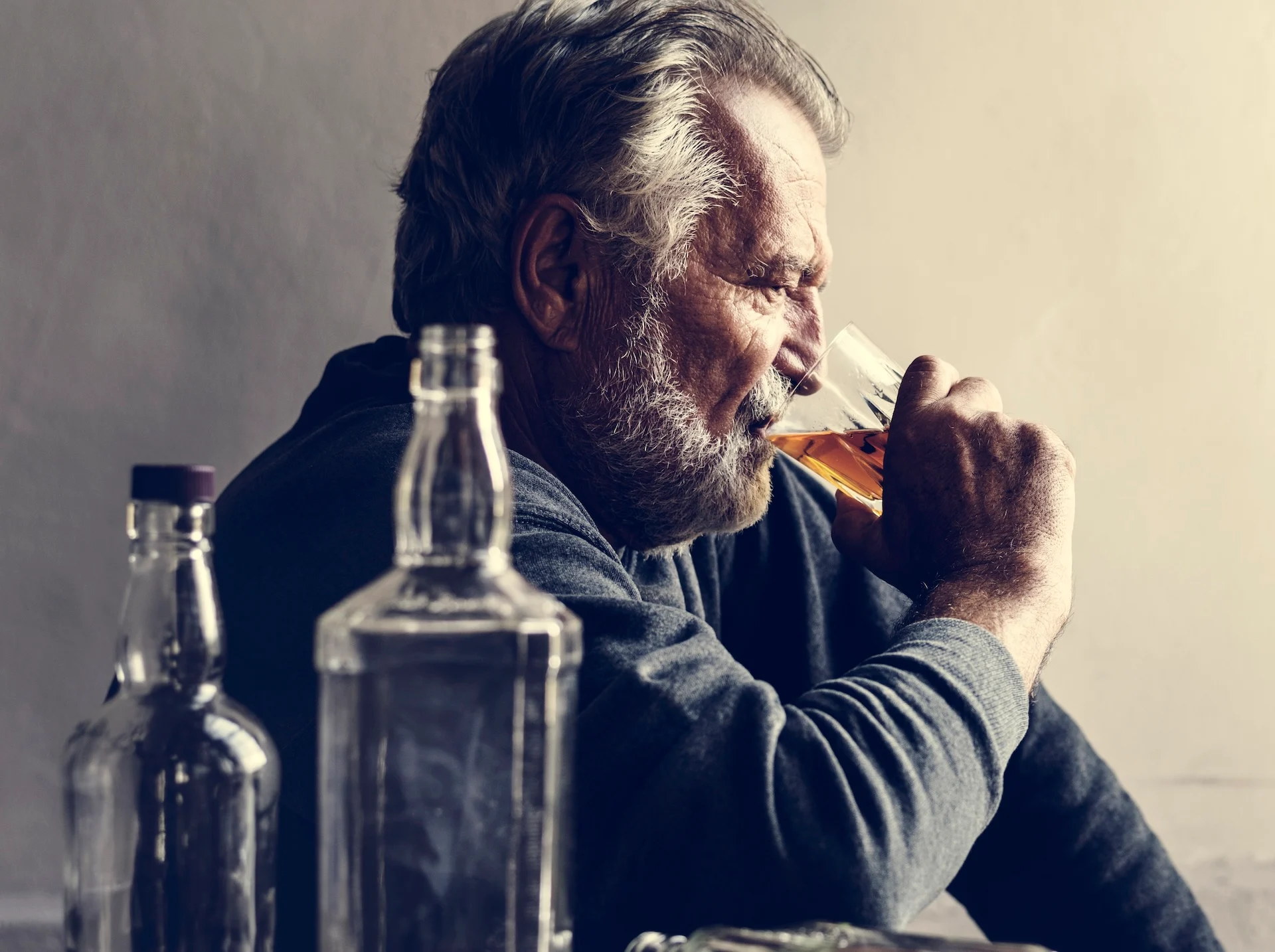 Is alcoholism genetic concept image showing an older man drinking whiskey alone at a table, surrounded by empty liquor bottles, suggesting family and biological risk for alcohol addiction.