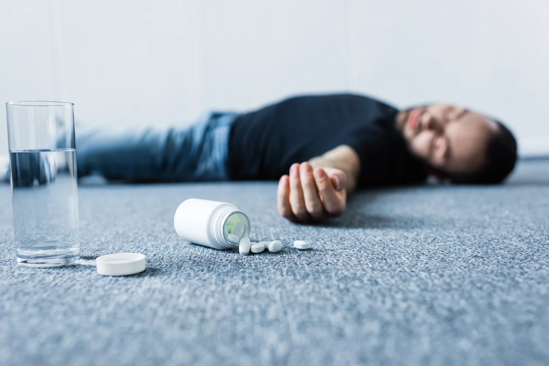 Unconscious man lying on the floor with a spilled pill bottle and glass of water in the foreground, illustrating the danger of a possible gabapentin overdose.